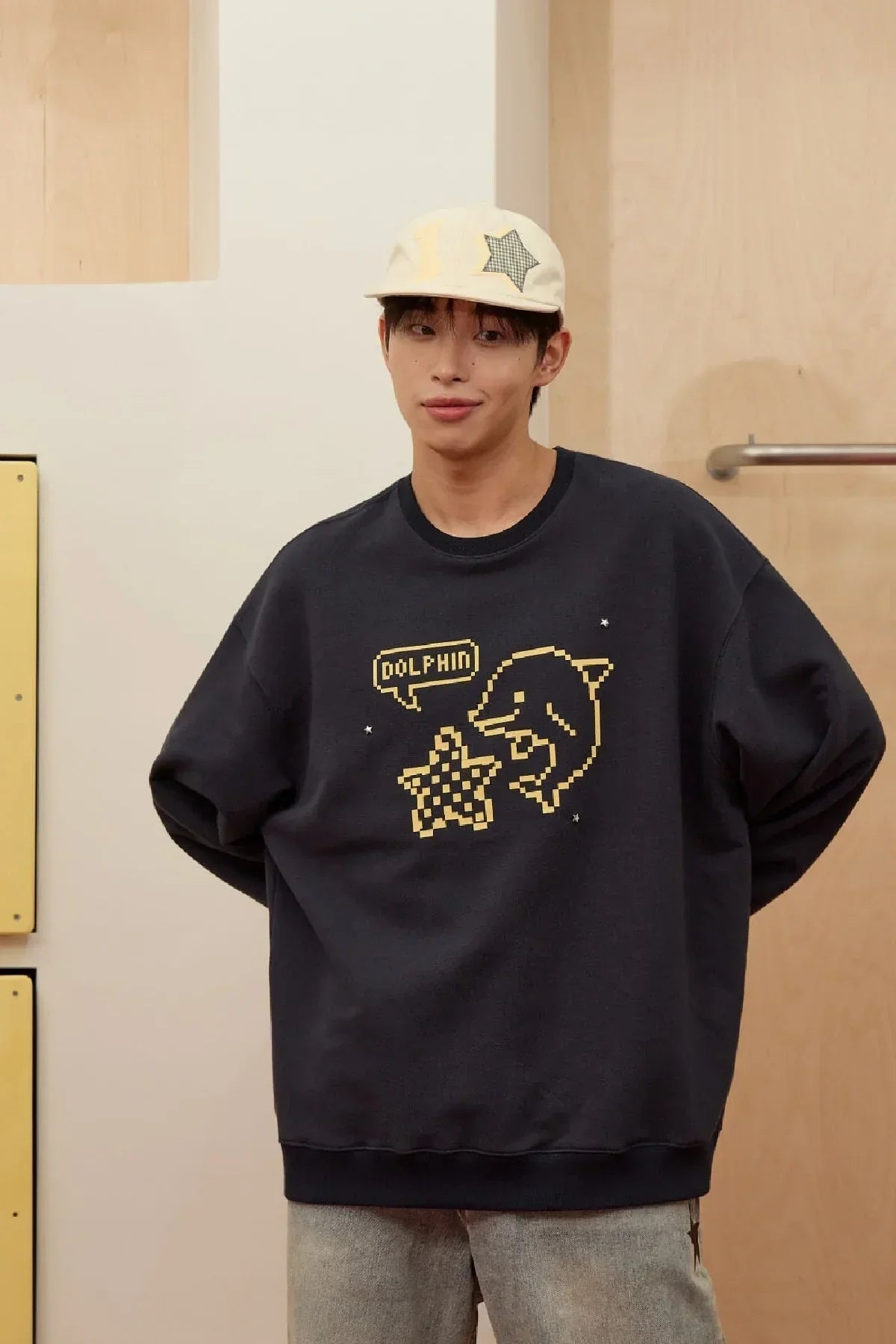 Man wearing navy sweatshirt with pixel dolphin and star design, paired with beige cap and light jeans