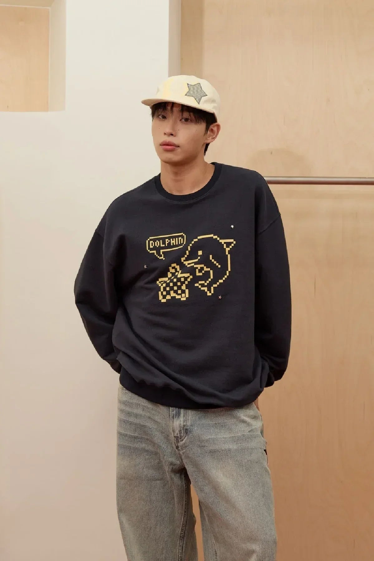 Man wearing navy sweatshirt with pixel art dolphin and star design, paired with light jeans and a beige star cap