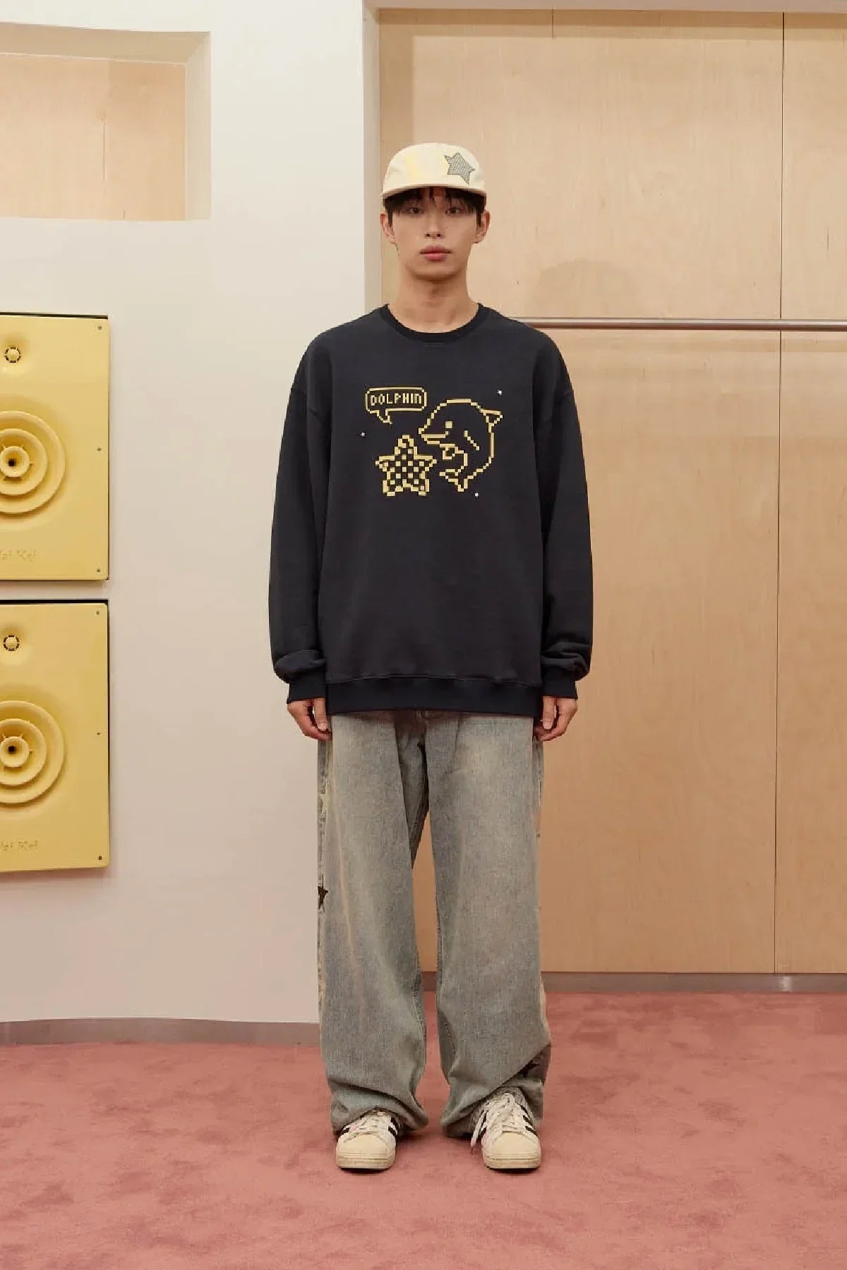 Young man wearing navy sweatshirt with pixel dolphin and yellow star design, beige cap, wide gray pants, and sneakers