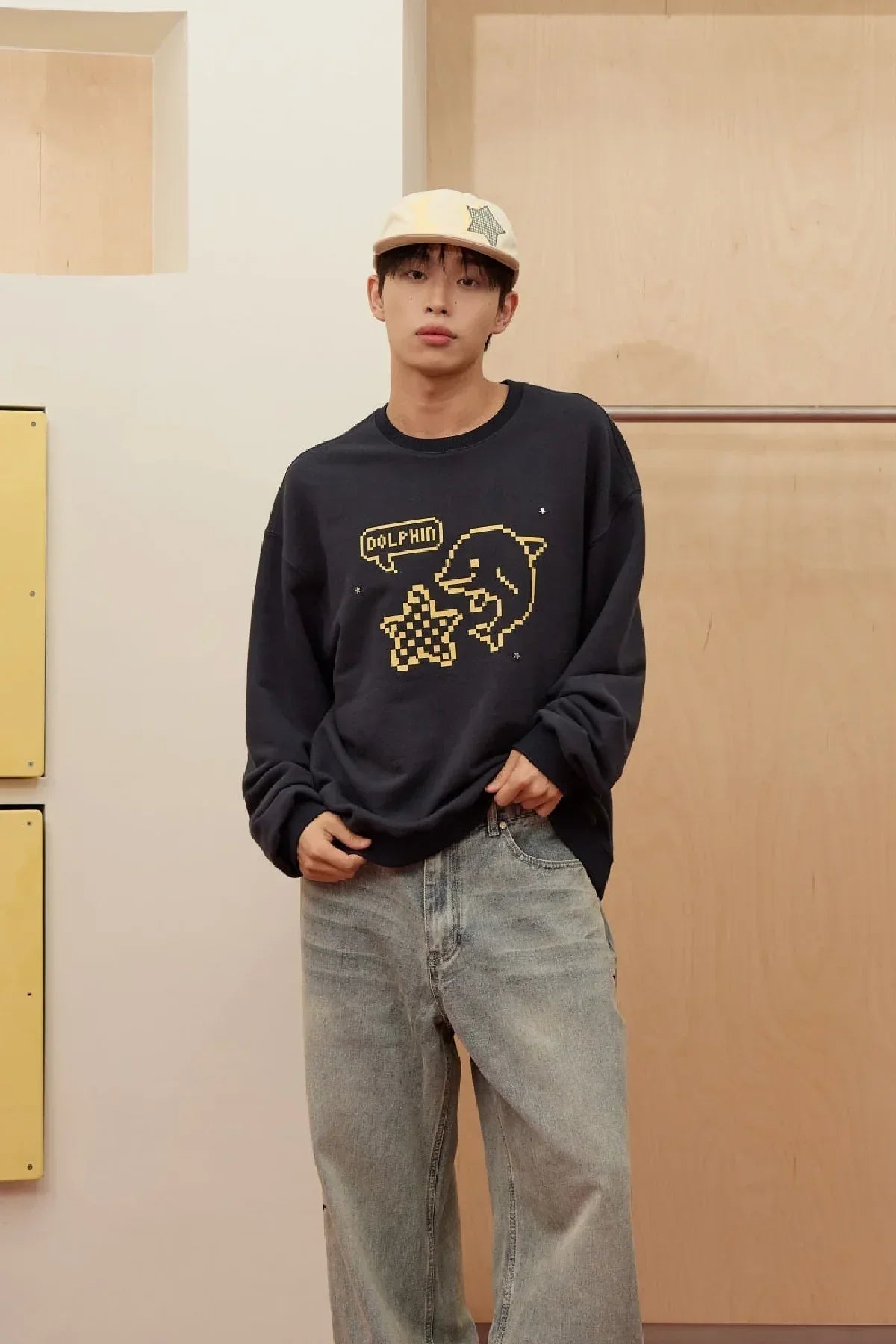 Man wearing navy sweatshirt with pixel dolphin and star design paired with light blue jeans and beige cap