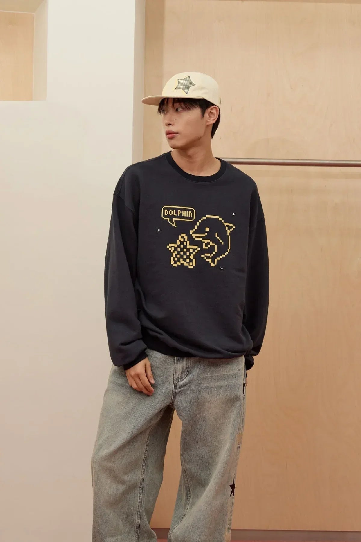 Man wearing navy sweatshirt with pixel art dolphin and star design, paired with light jeans and beige cap
