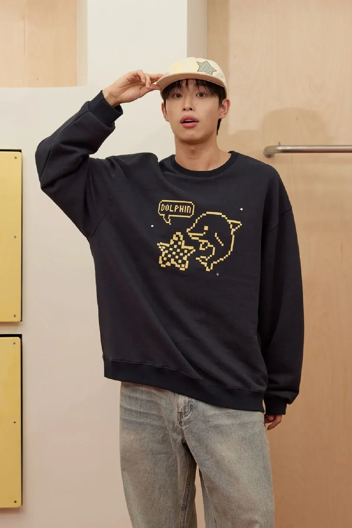 Young man wearing navy sweatshirt with pixel dolphin and star design, paired with light jeans and beige star cap