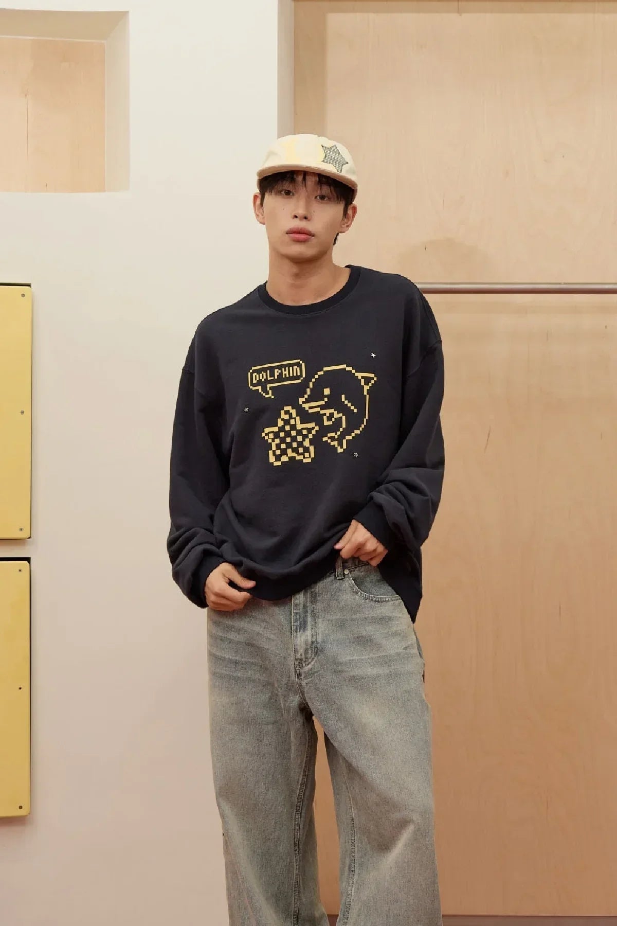 Man wearing navy sweatshirt with yellow pixel dolphin and star design, beige cap, and light denim jeans