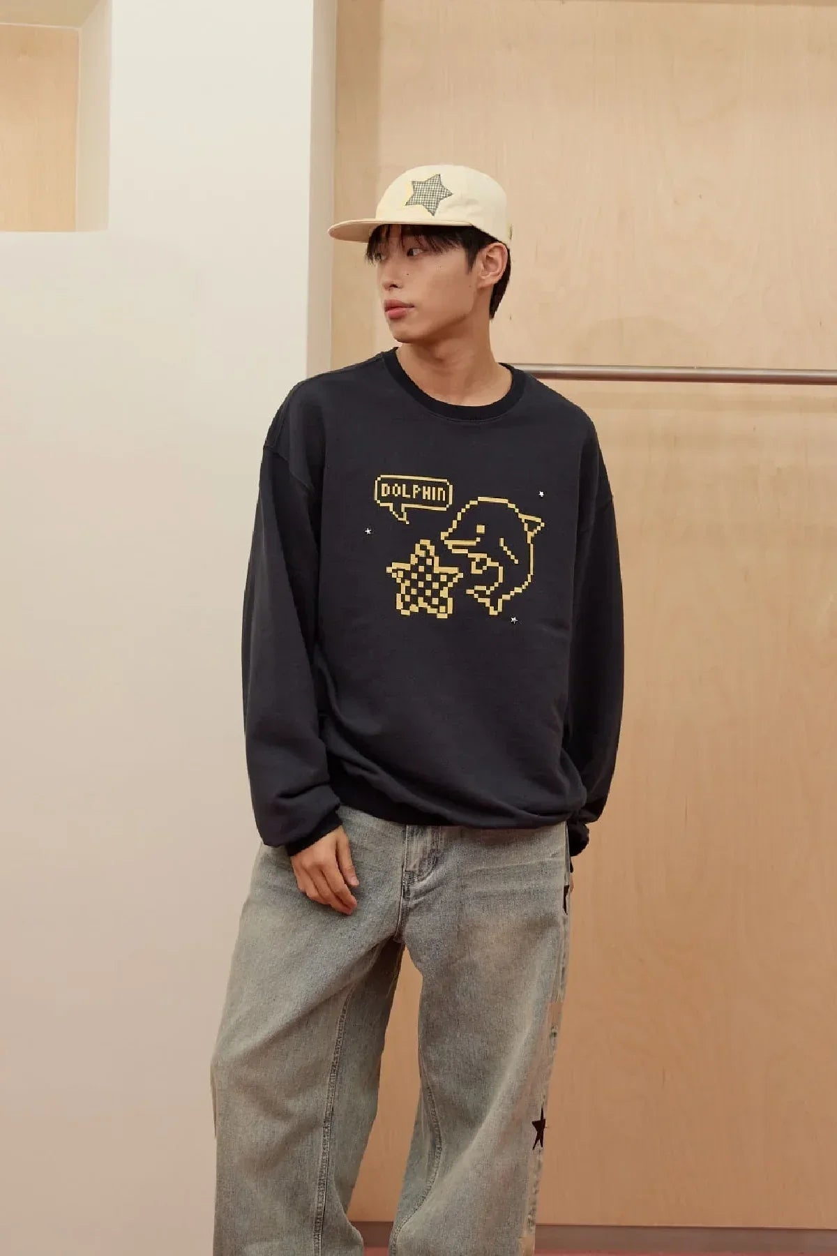 Man wearing navy sweatshirt with pixel dolphin and star graphic, cream cap with star, and loose jeans