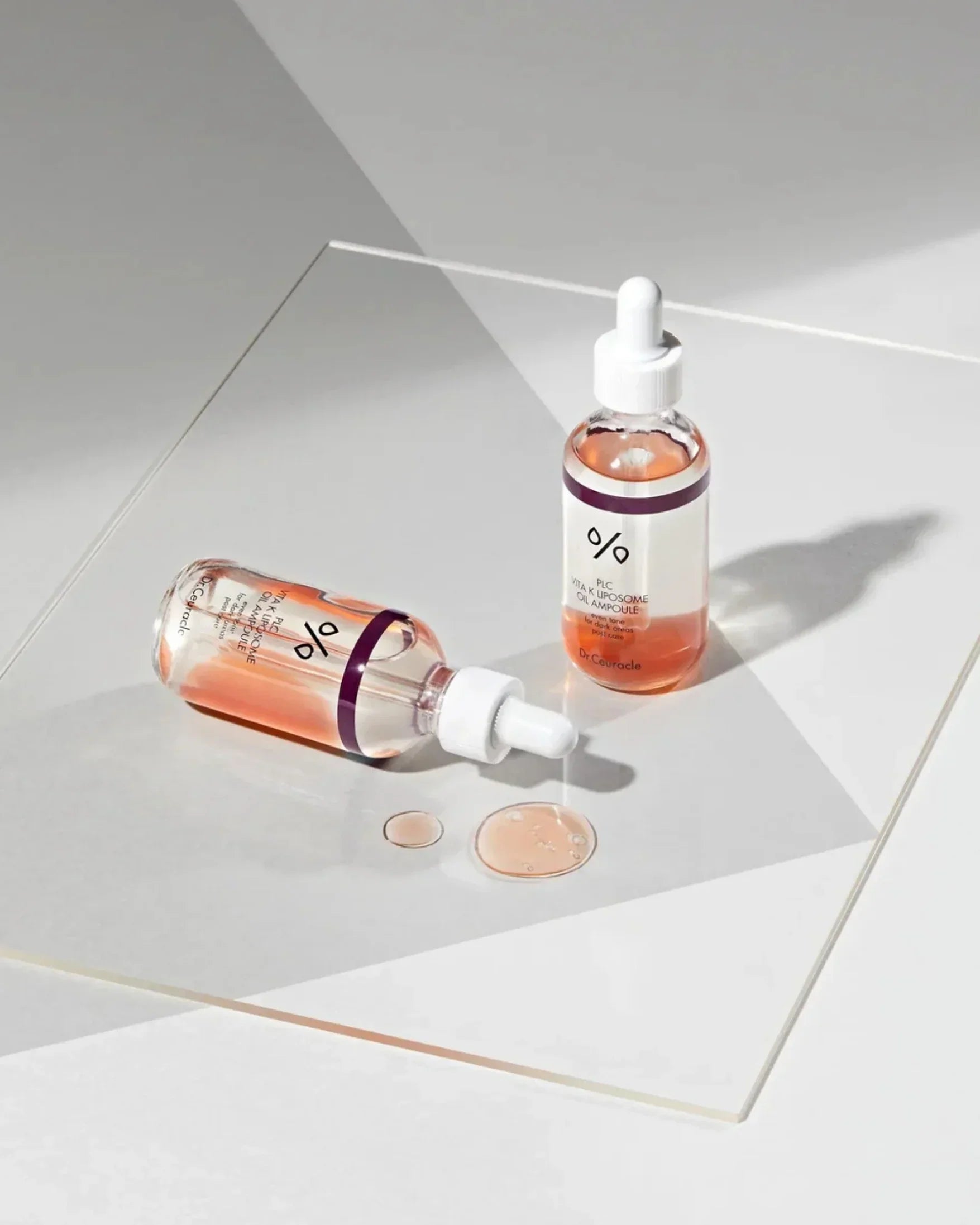 Two bottles of PLC Vita K Liposome Oil Ampoule with dropper and drops on white surface