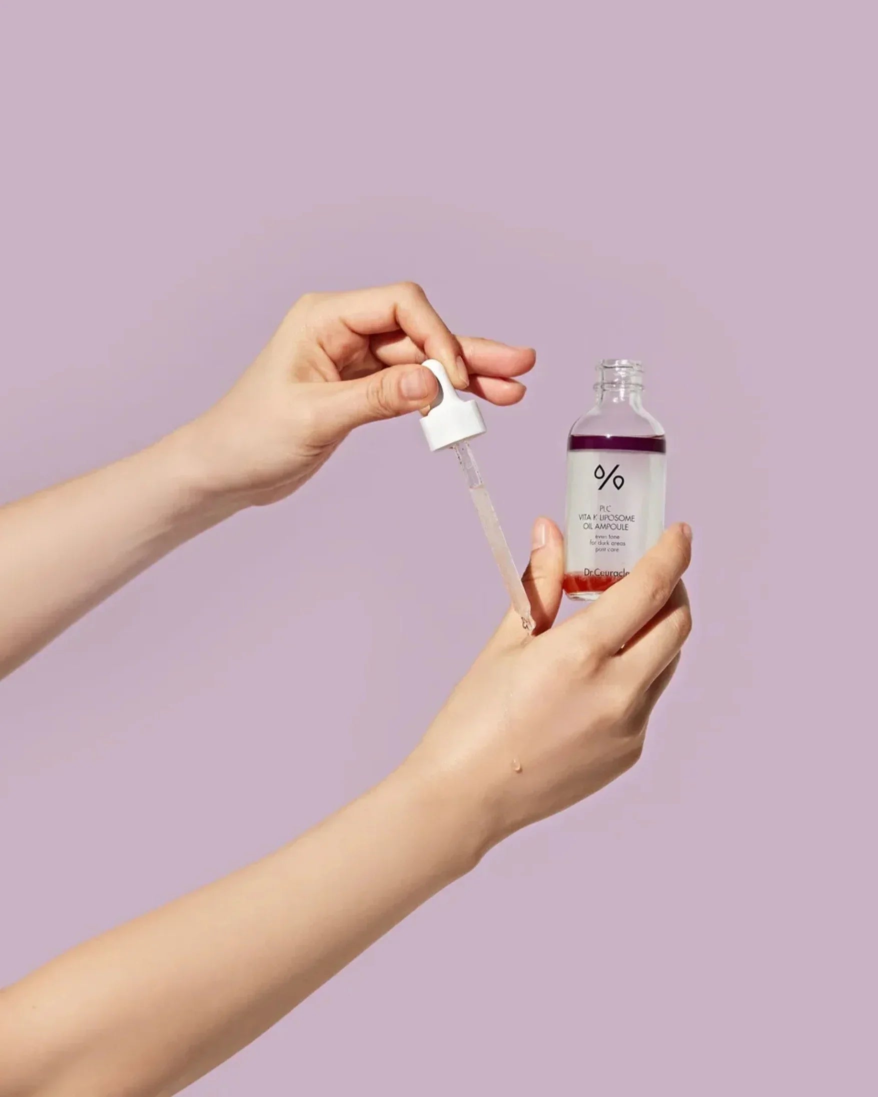 Hands holding and applying Dr. Ceuracle PLC Vita K Liposome Oil Ampoule serum with dropper on light purple background