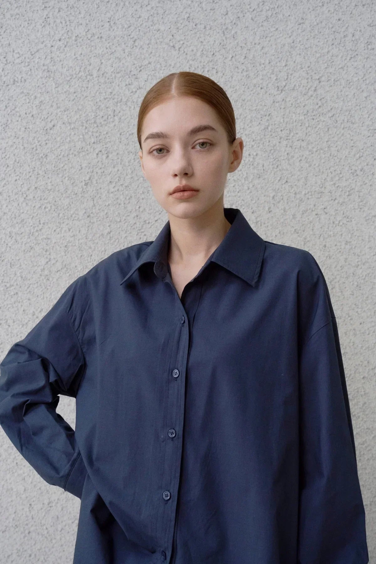 Young woman wearing a navy poplin button-up shirt with a textured gray wall background