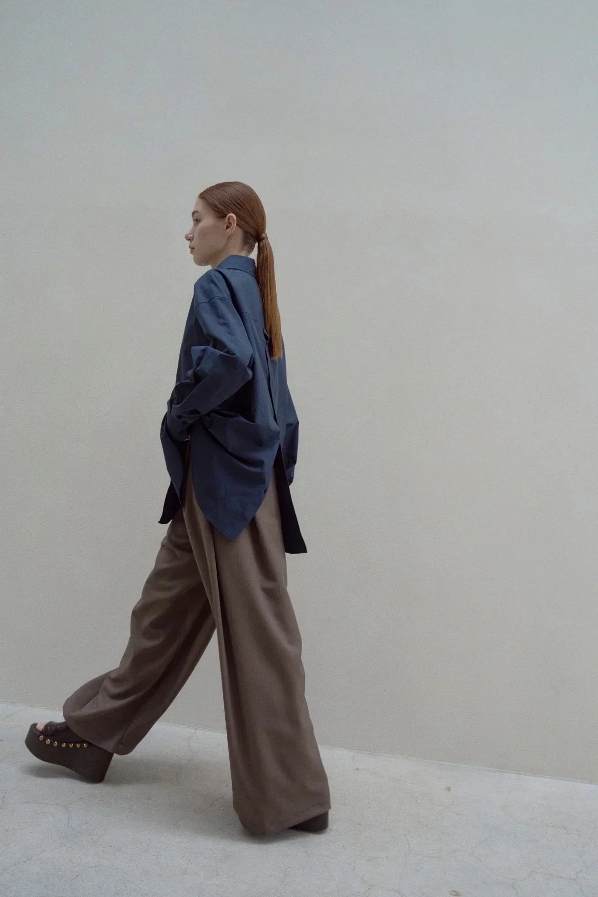 Woman wearing navy poplin back button shirt and wide brown trousers walking against plain wall