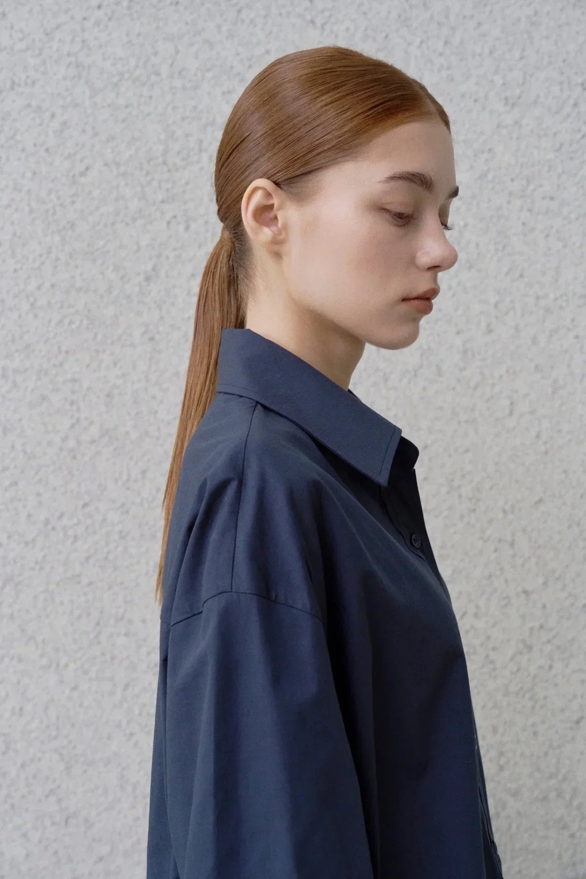 Profile of woman with slicked-back hair wearing navy oversized button-up shirt against textured light gray wall