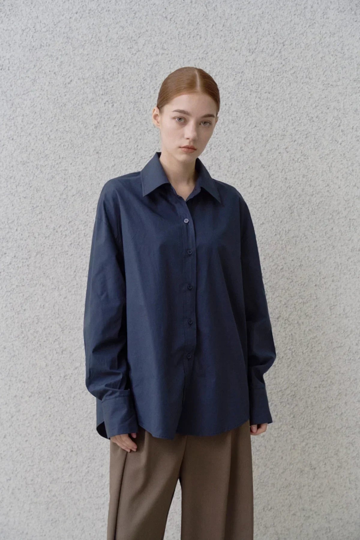 Woman wearing a navy blue oversized button-up poplin shirt with brown wide-leg pants against a light textured wall