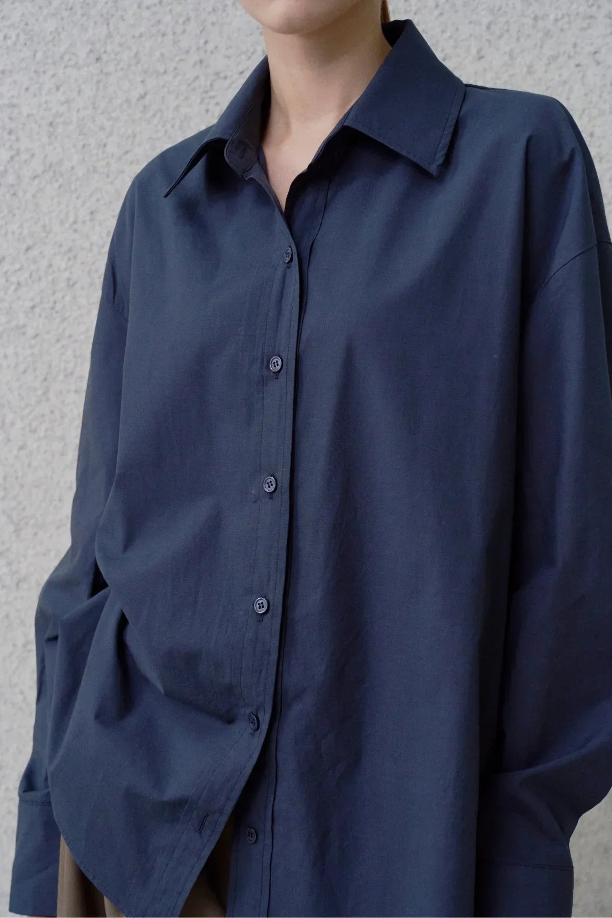 Close-up of navy blue poplin shirt with buttons and wide collar worn by model against textured wall