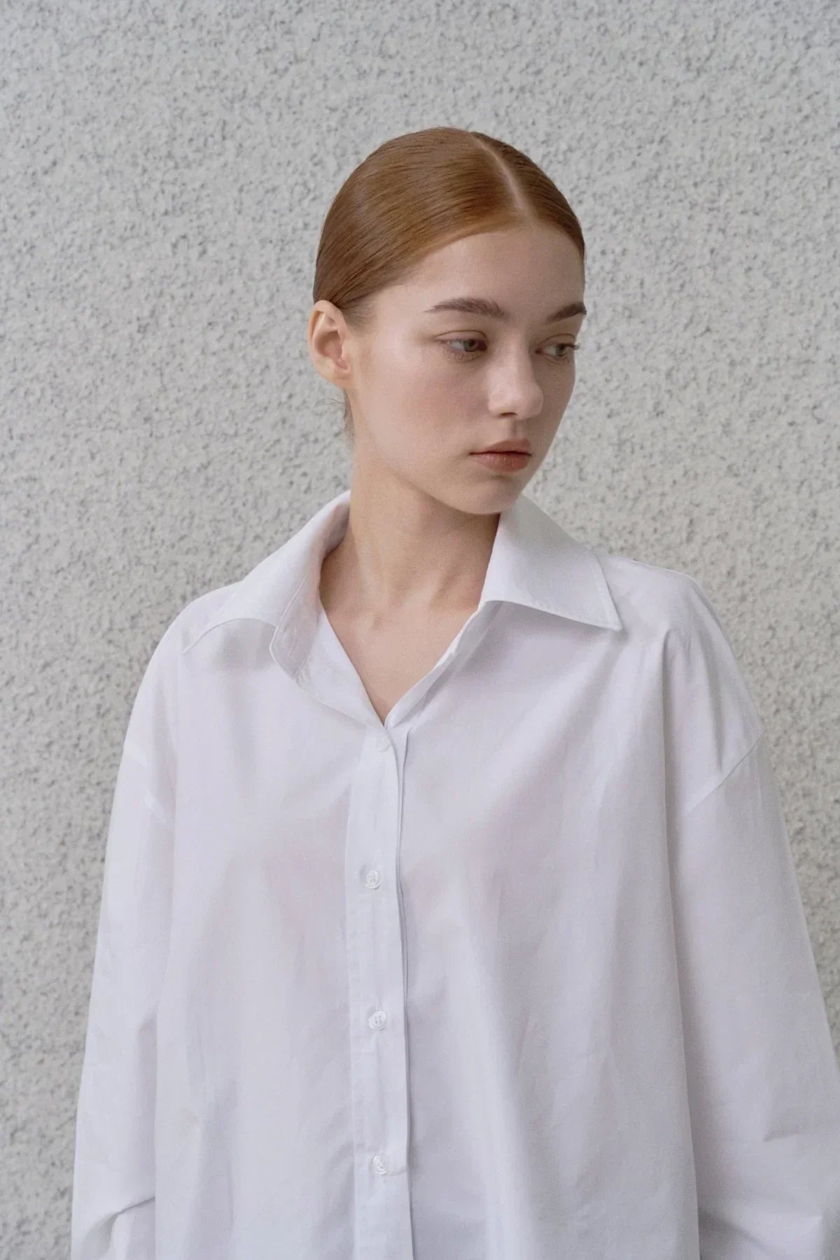 Young woman with red hair wearing a white poplin button-down shirt against a textured gray wall