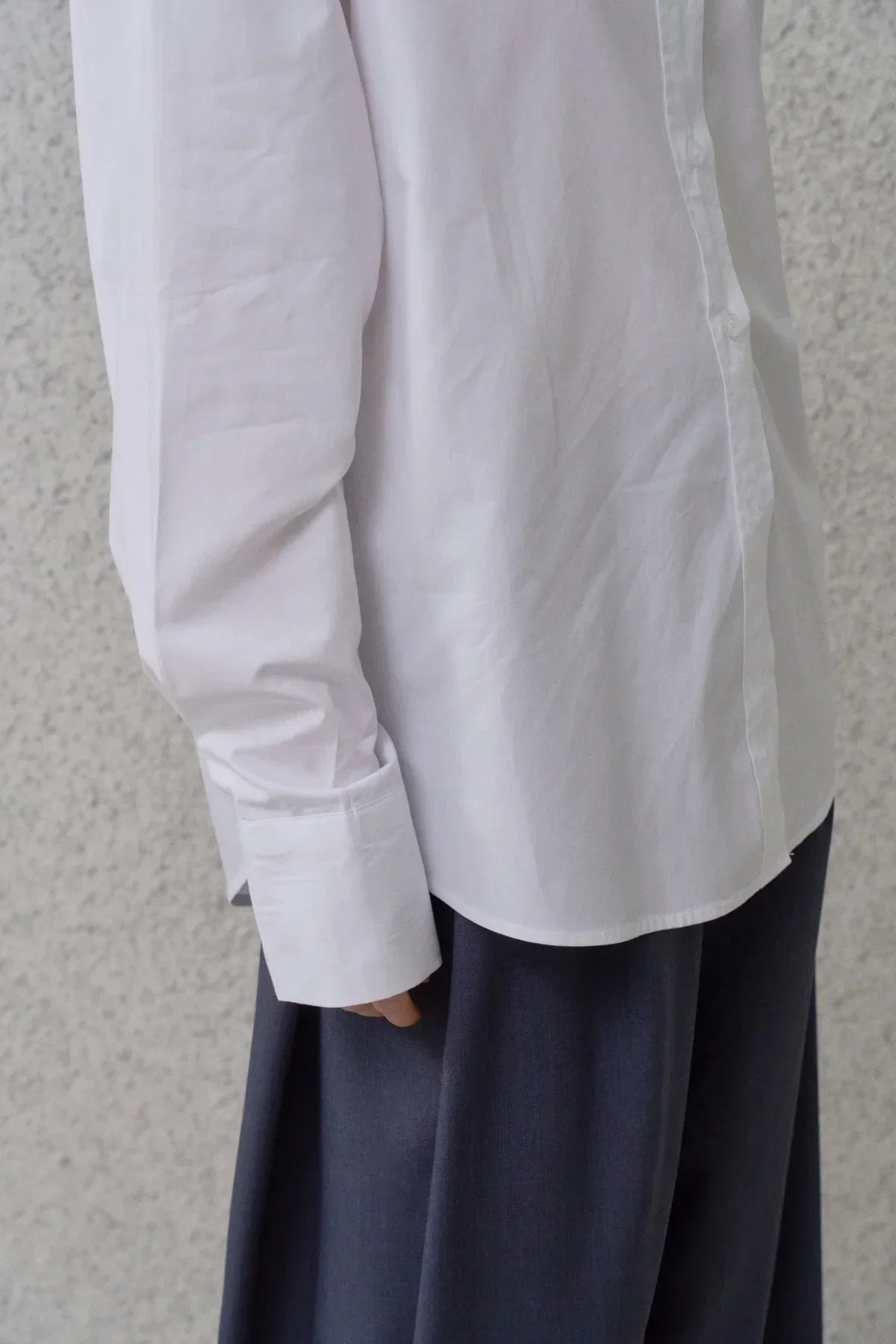 Close-up of white poplin shirt cuff and button placket paired with dark pants against textured wall