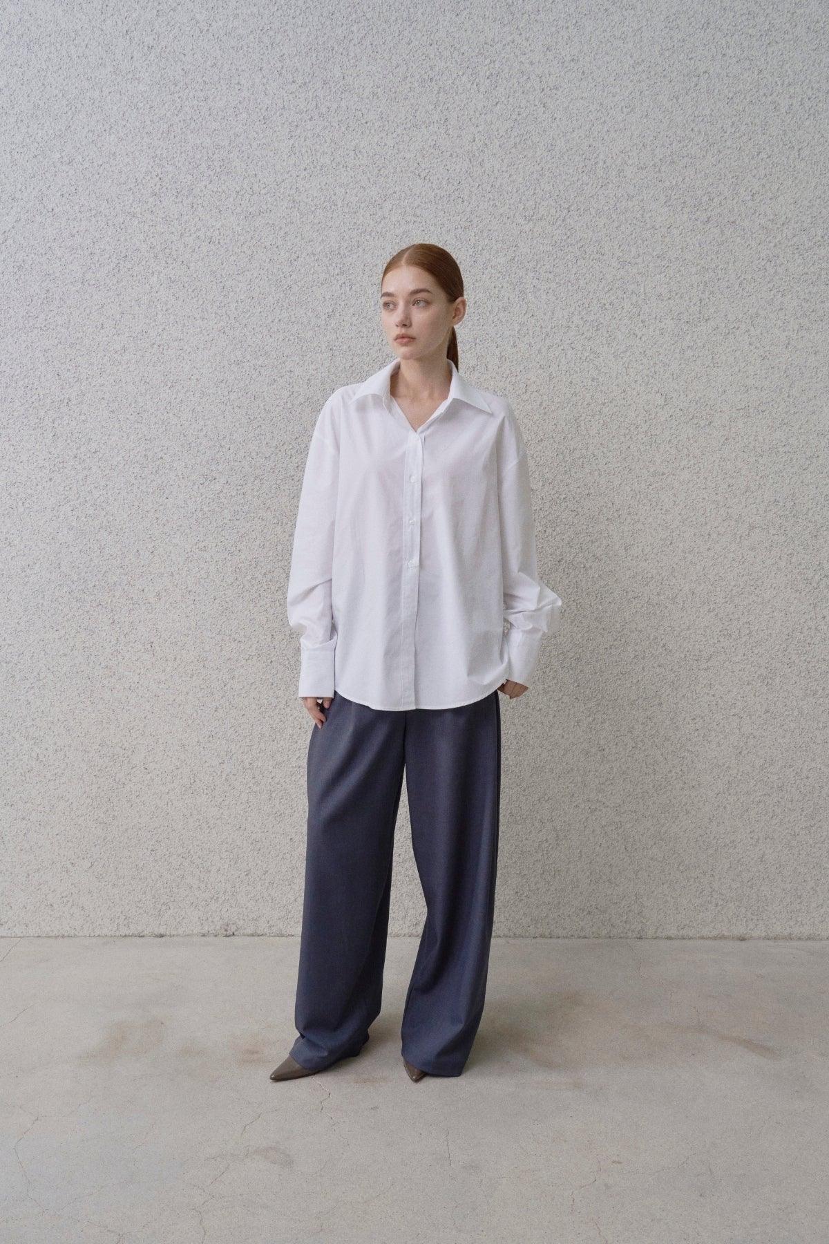 Woman wearing white poplin shirt and loose dark wide-leg pants standing against textured wall