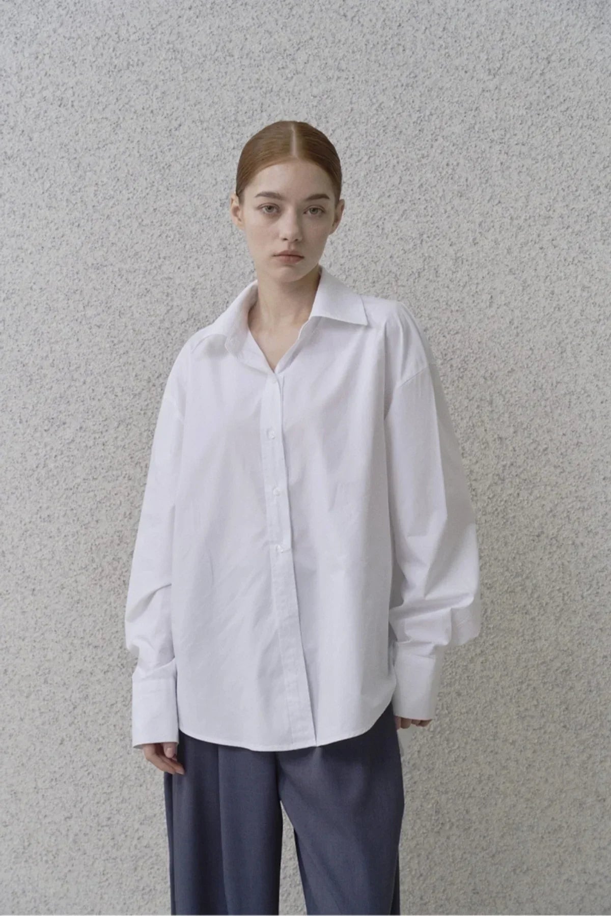 Woman wearing a loose white poplin shirt and gray trousers against a textured wall