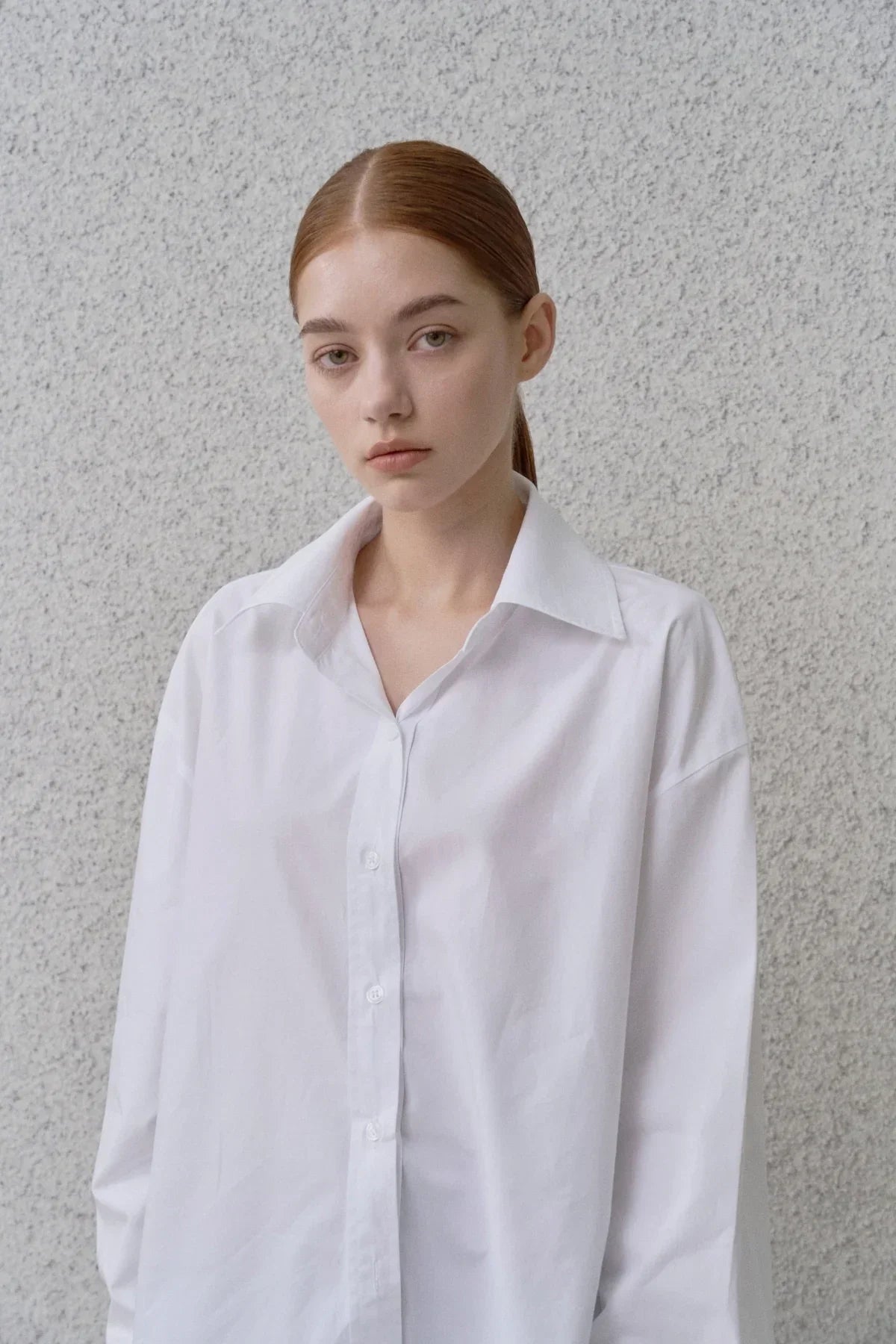 Young woman with red hair wearing a white oversized poplin button-up shirt against a textured gray wall