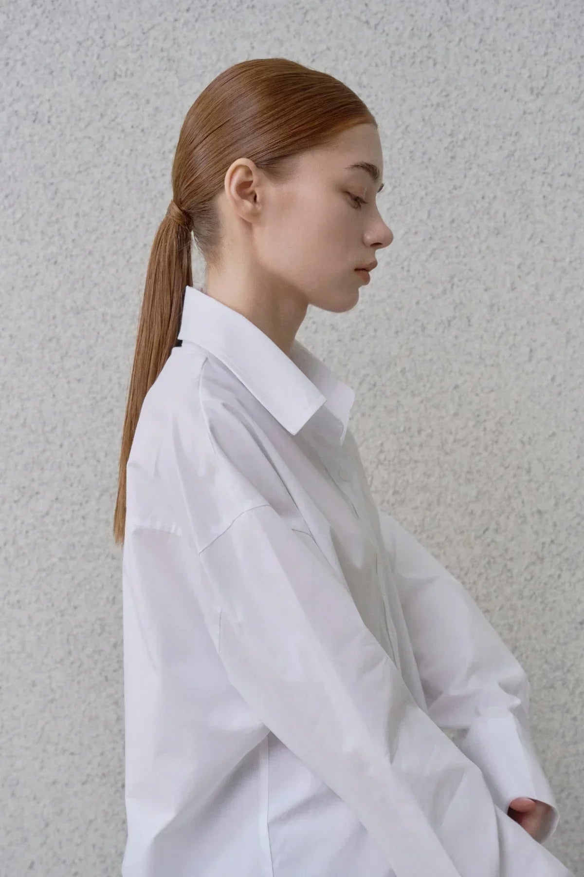Side profile of a woman with straight brown hair tied back, wearing an oversized white button-up shirt against a textured gray wall