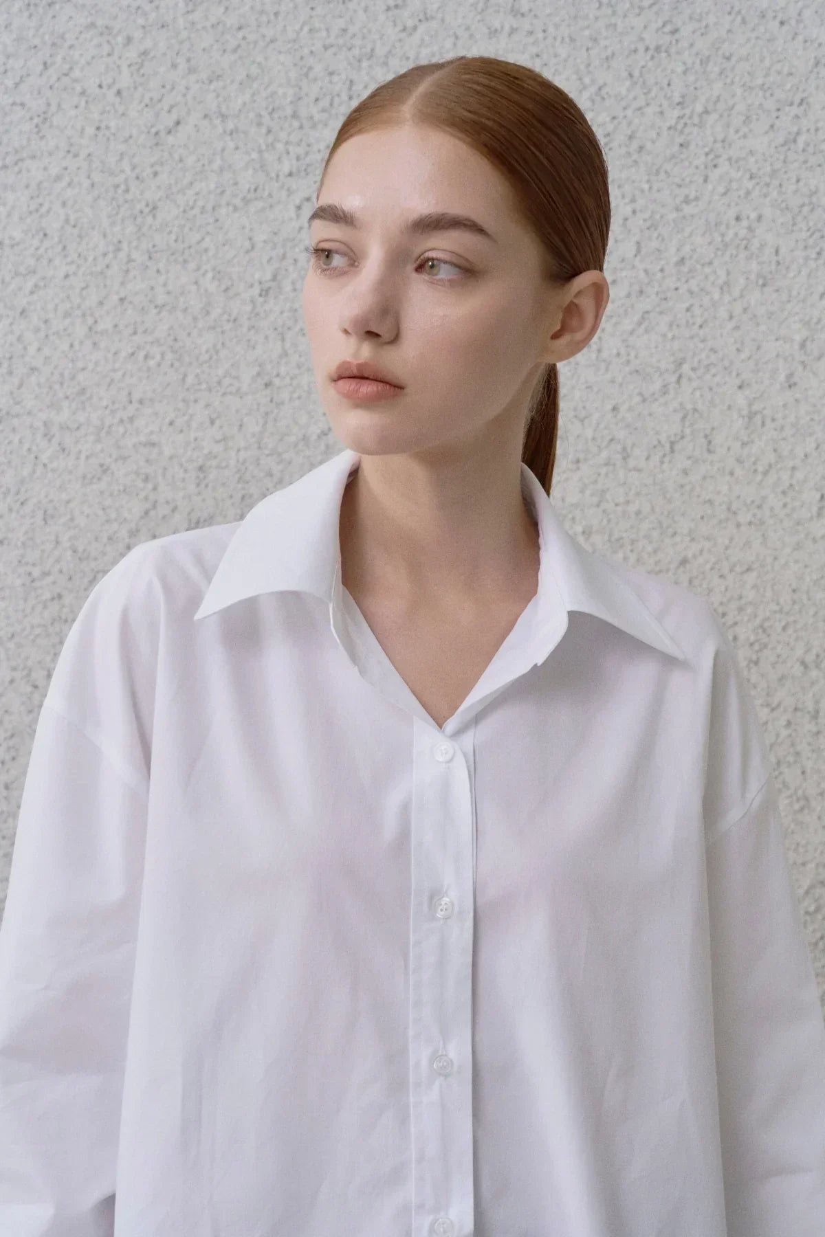 Young woman with red hair in a sleek ponytail wearing a white button-up shirt against a textured gray wall