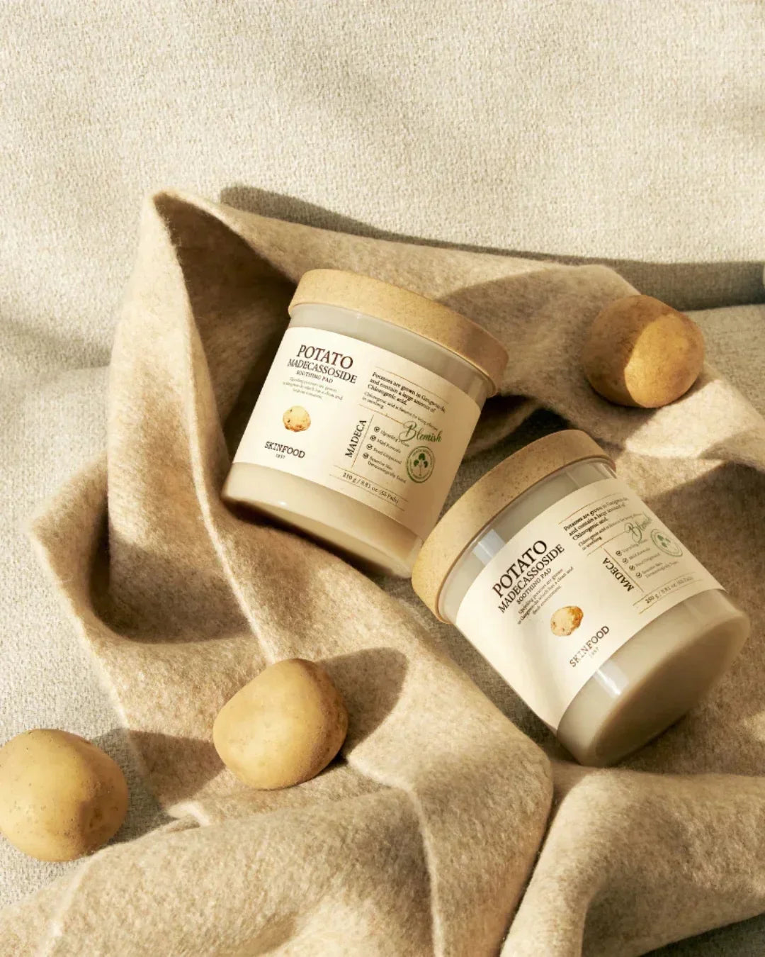 Two jars of Skinfood Potato Madecassoside Soothing Pad on beige fabric with whole potatoes