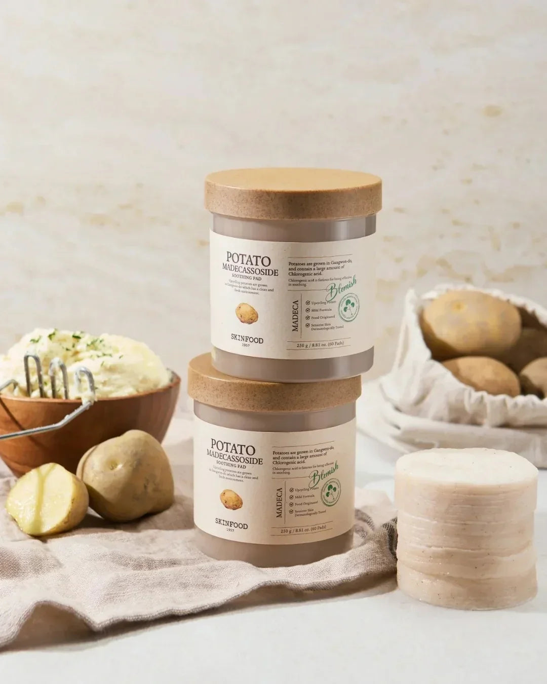 Two jars of Potato Madecassoside Soothing Pads by Skinfood stacked with sliced and whole potatoes, mashed potatoes, and pads on a beige cloth
