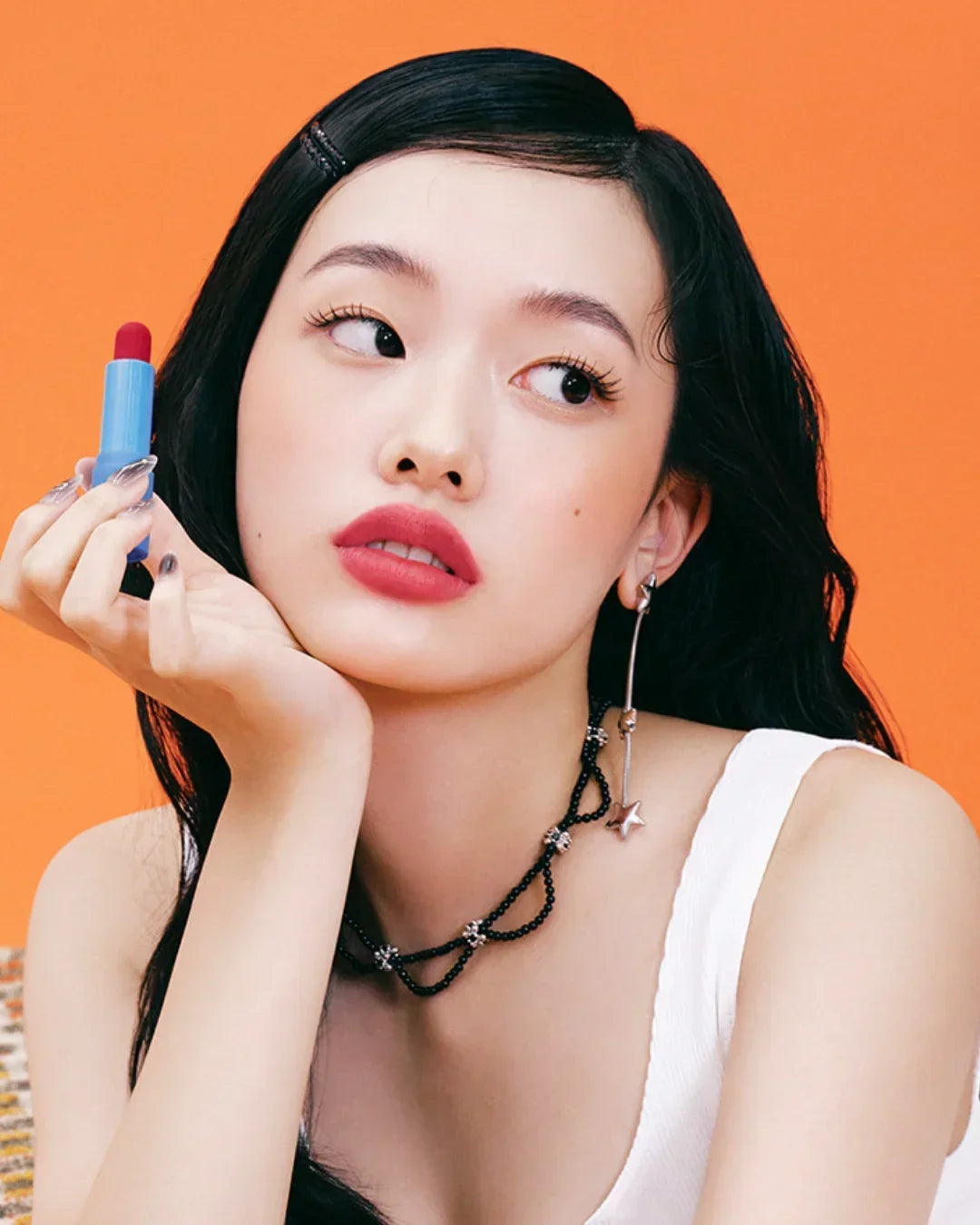 Young woman with dark hair holding red lip balm in blue tube against orange background, wearing white top and black necklace