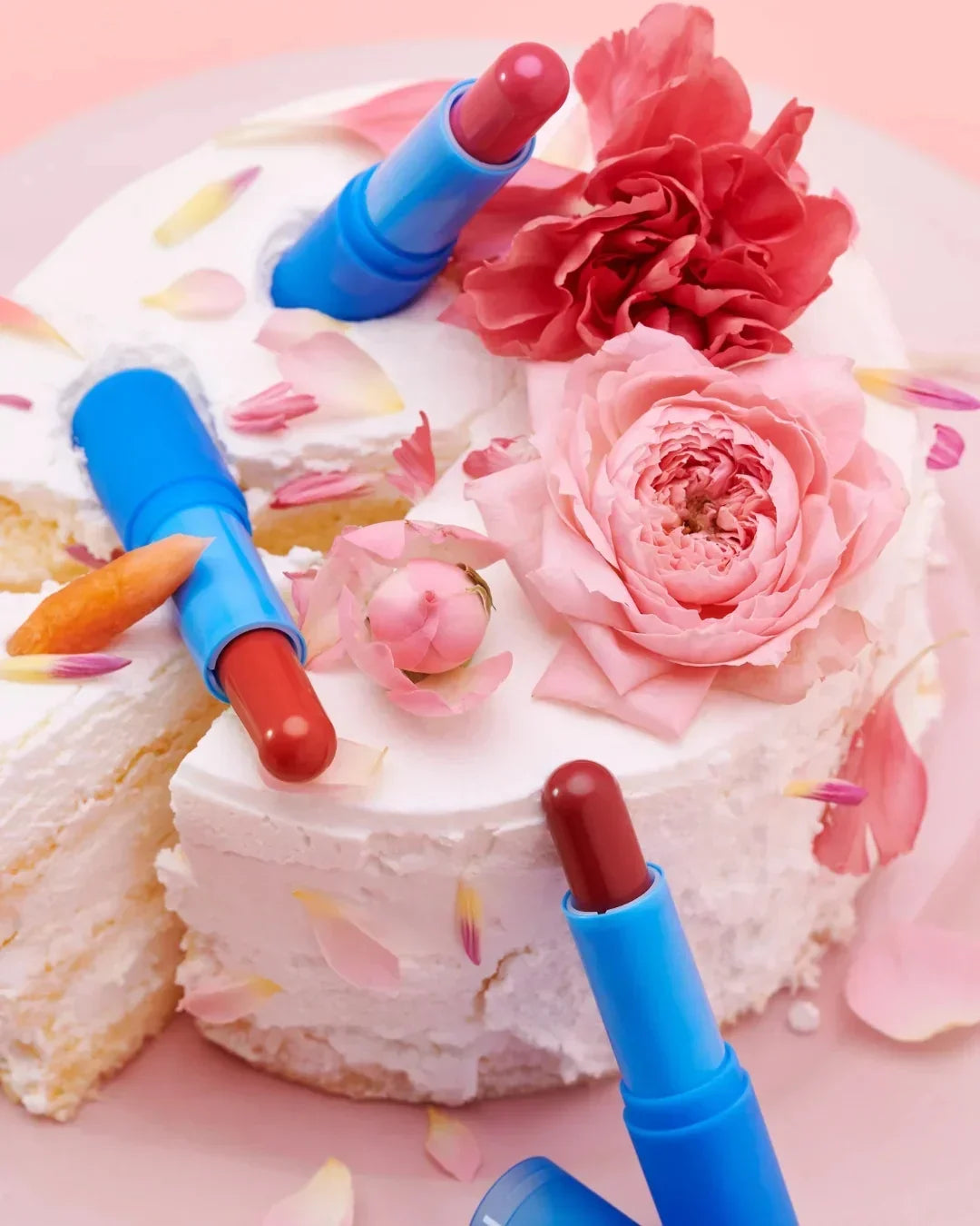 White frosted cake decorated with pink flowers and multiple shades of red lipstick in blue tubes