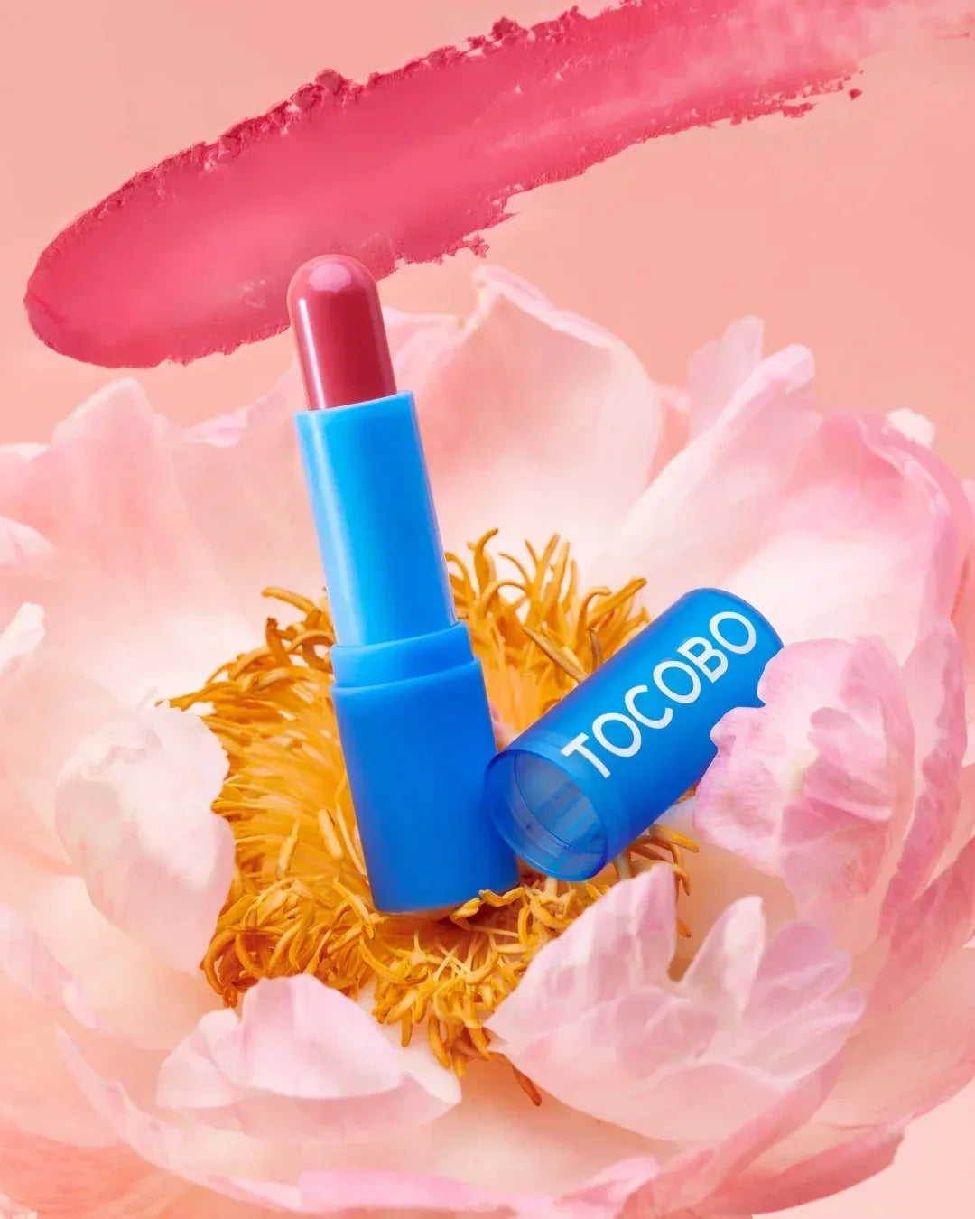 TOCOBO pink lip balm in blue tube on pink flower with pink lipstick smear background