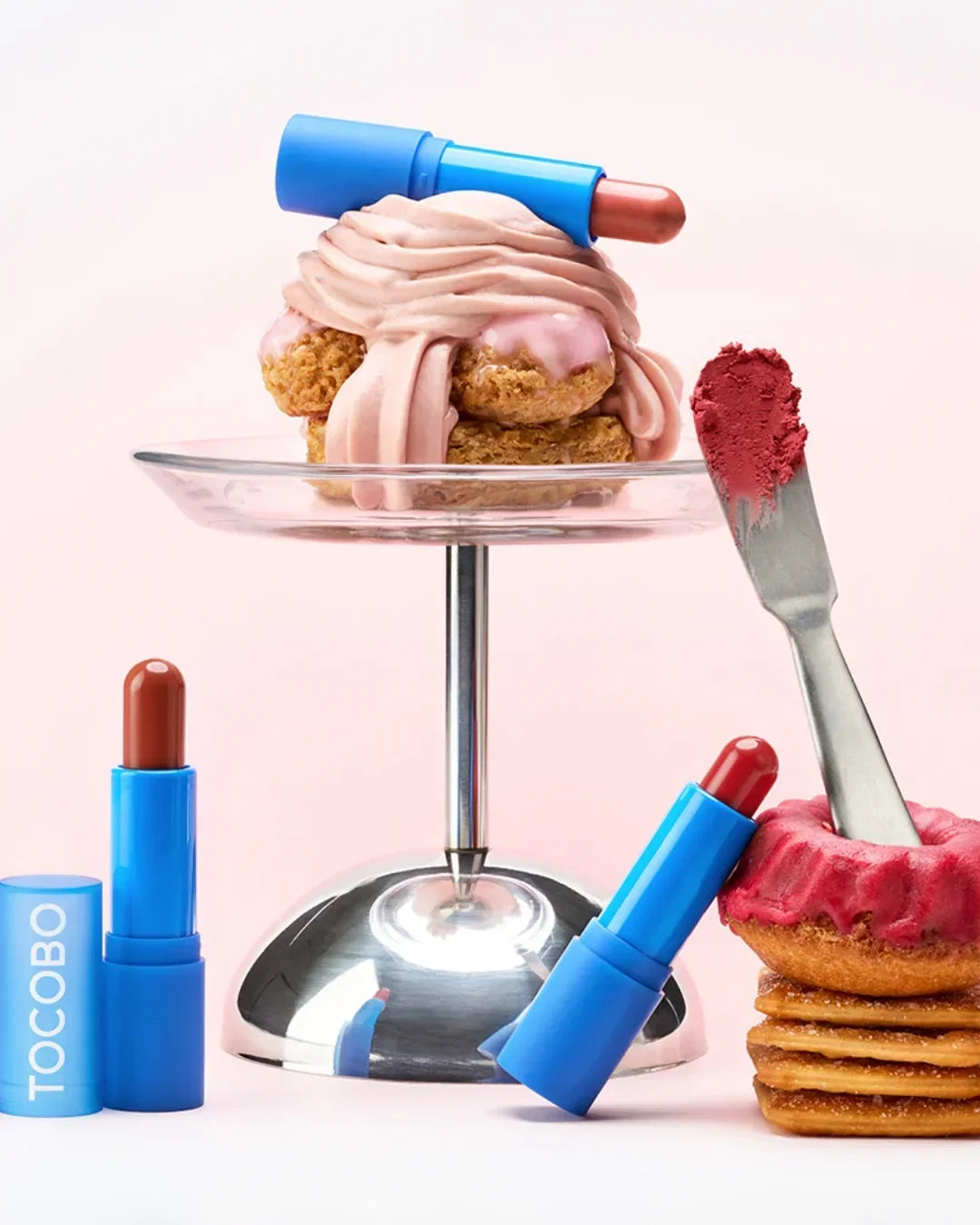 Blue Tocobo lip balms beside and on top of stacked pastries with pink and red frosting, pink background
