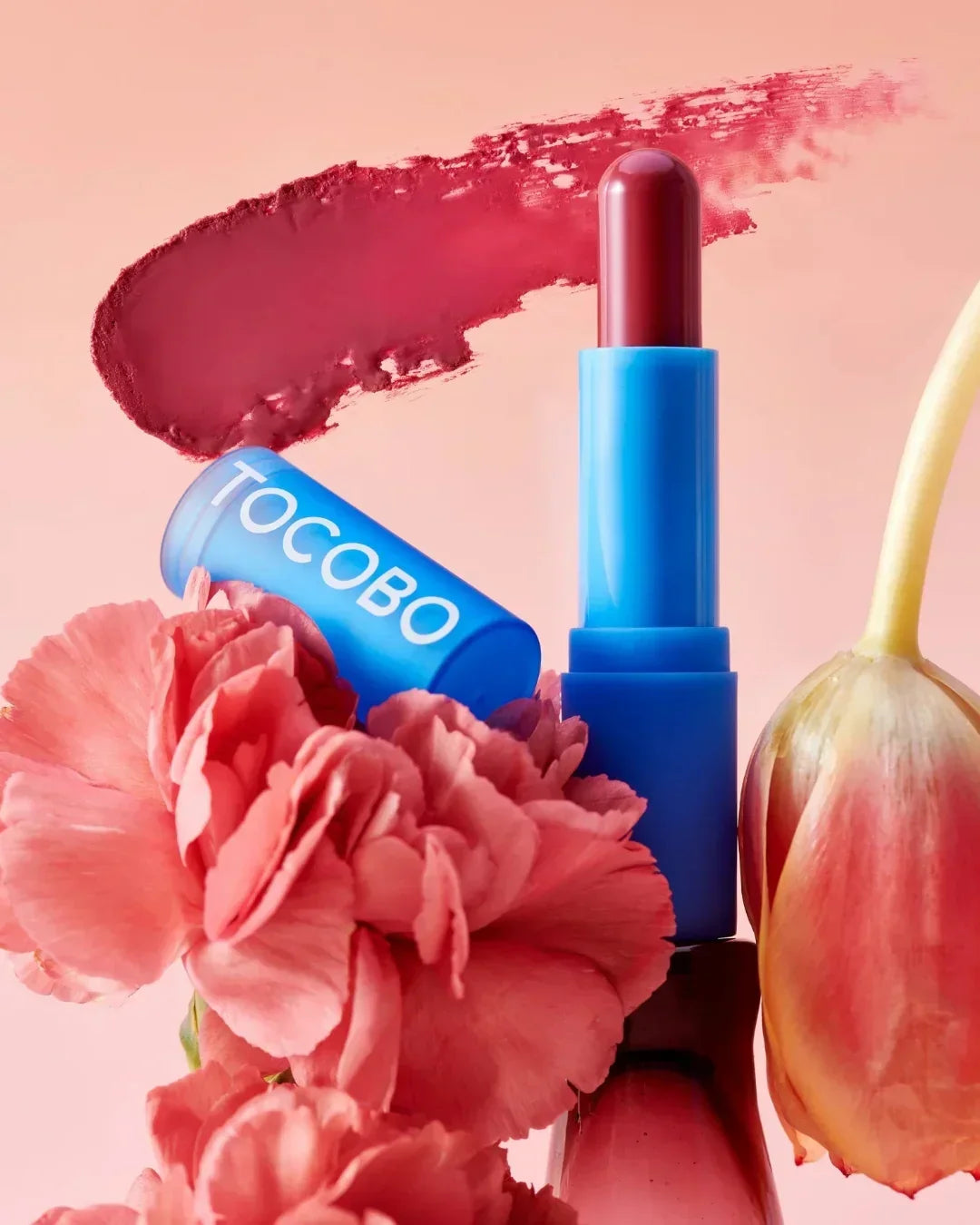 Tocobo red lipstick with blue case surrounded by pink carnation and tulip flowers on peach background