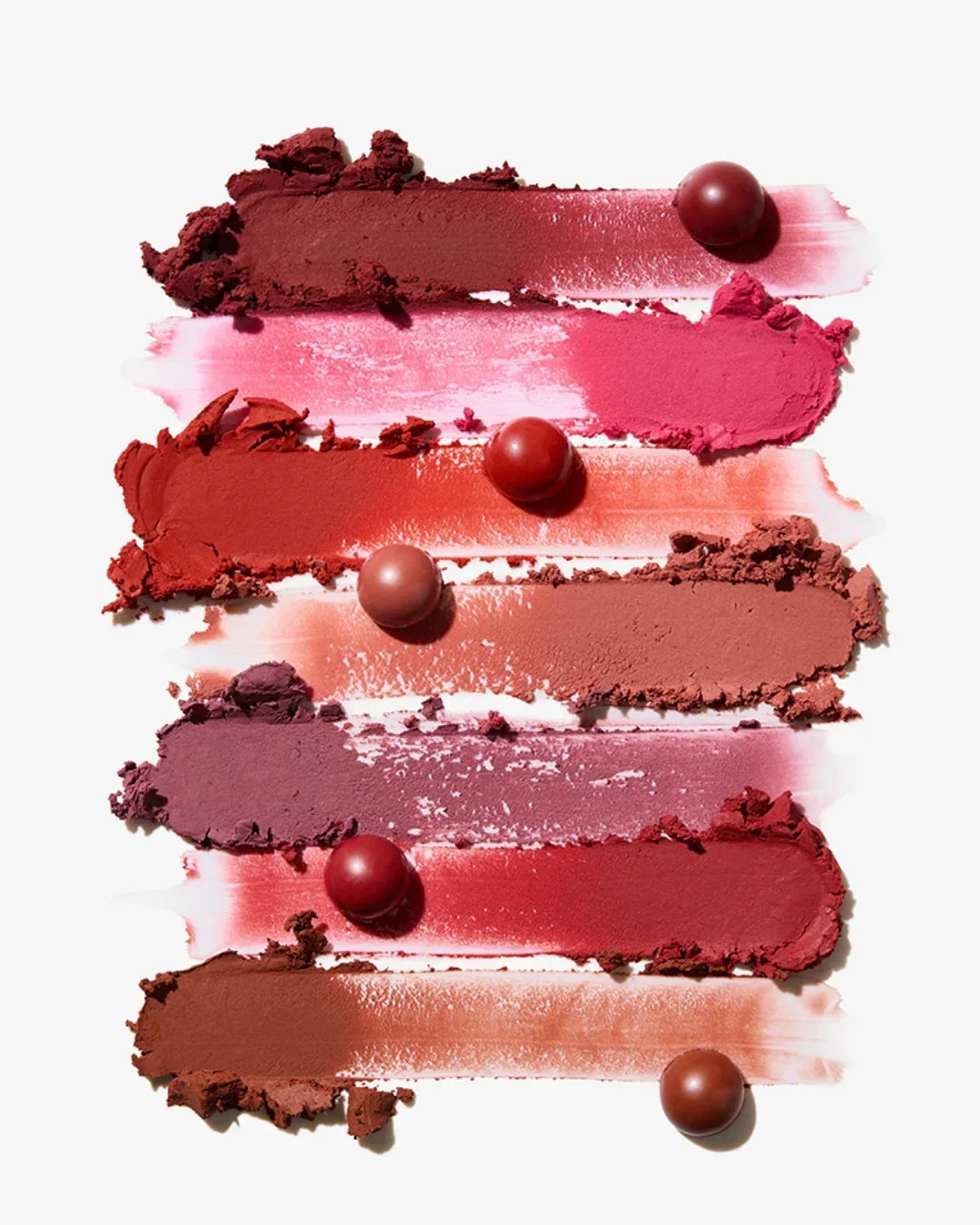 Smears of six powder lip balms in red, pink, mauve, and brown shades with matching round balm balls on white background