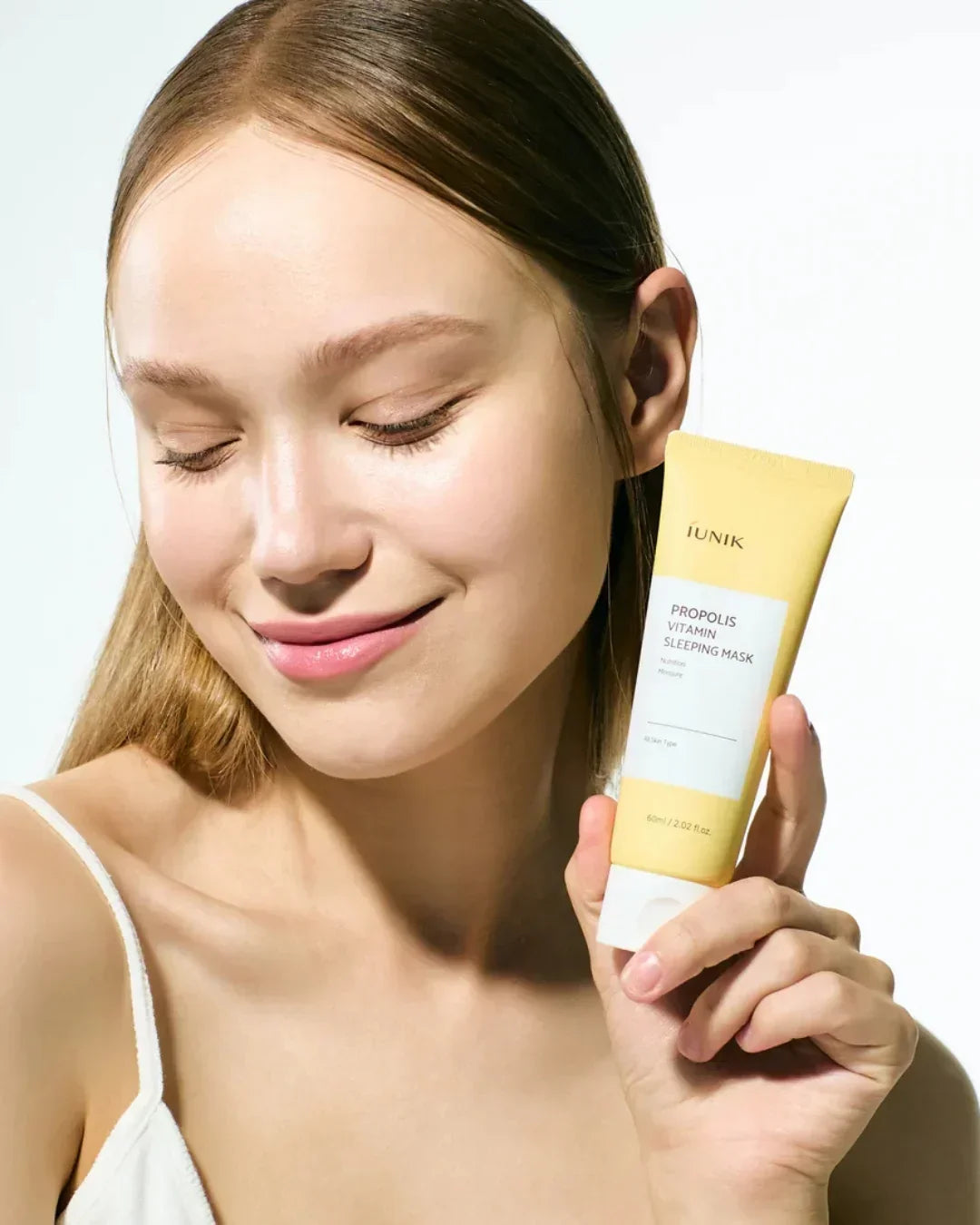 Smiling woman with clear skin holding iUNIK Propolis Vitamin Sleeping Mask tube against white background