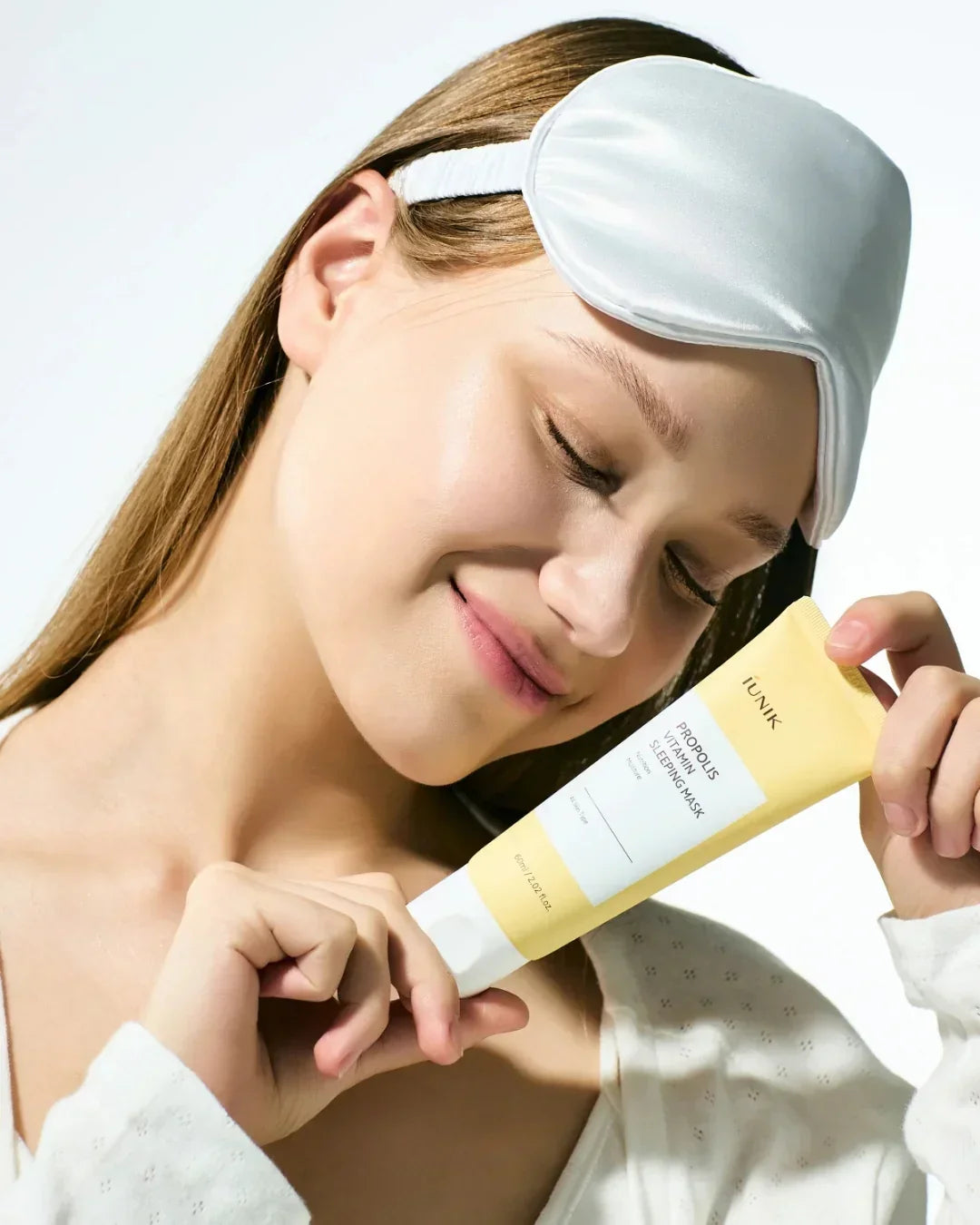 Smiling woman with satin sleep mask holds iUNIK Propolis Vitamin Sleeping Mask tube, skincare