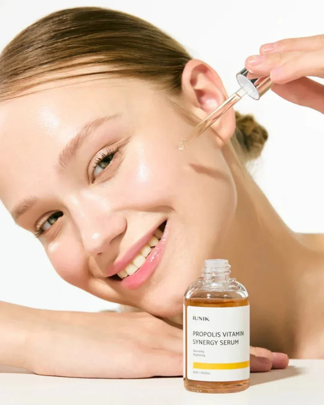 Smiling woman applying IUNIK Propolis Vitamin Synergy Serum with dropper on glowing skin