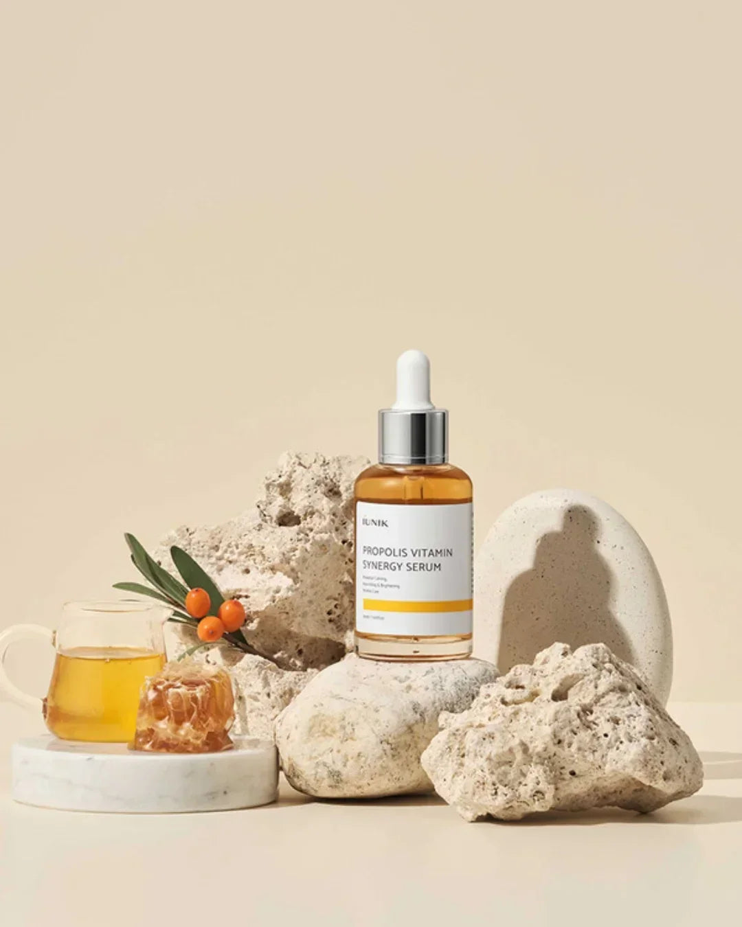 Iunik Propolis Vitamin Synergy Serum bottle on textured stones with honeycomb, tea, and sea buckthorn berries
