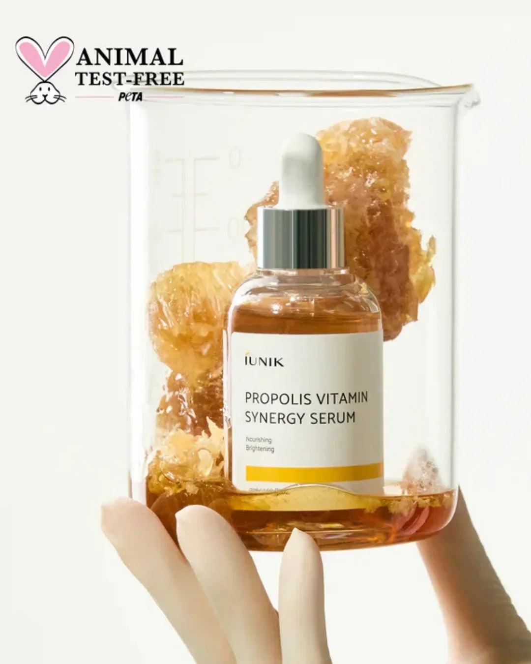 Hand holding IUNIK Propolis Vitamin Synergy Serum bottle inside beaker with propolis chunks, animal test-free logo