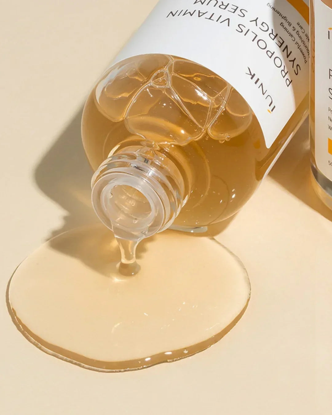 Close-up of Propolis Vitamin Synergy Serum pouring from clear bottle on beige surface