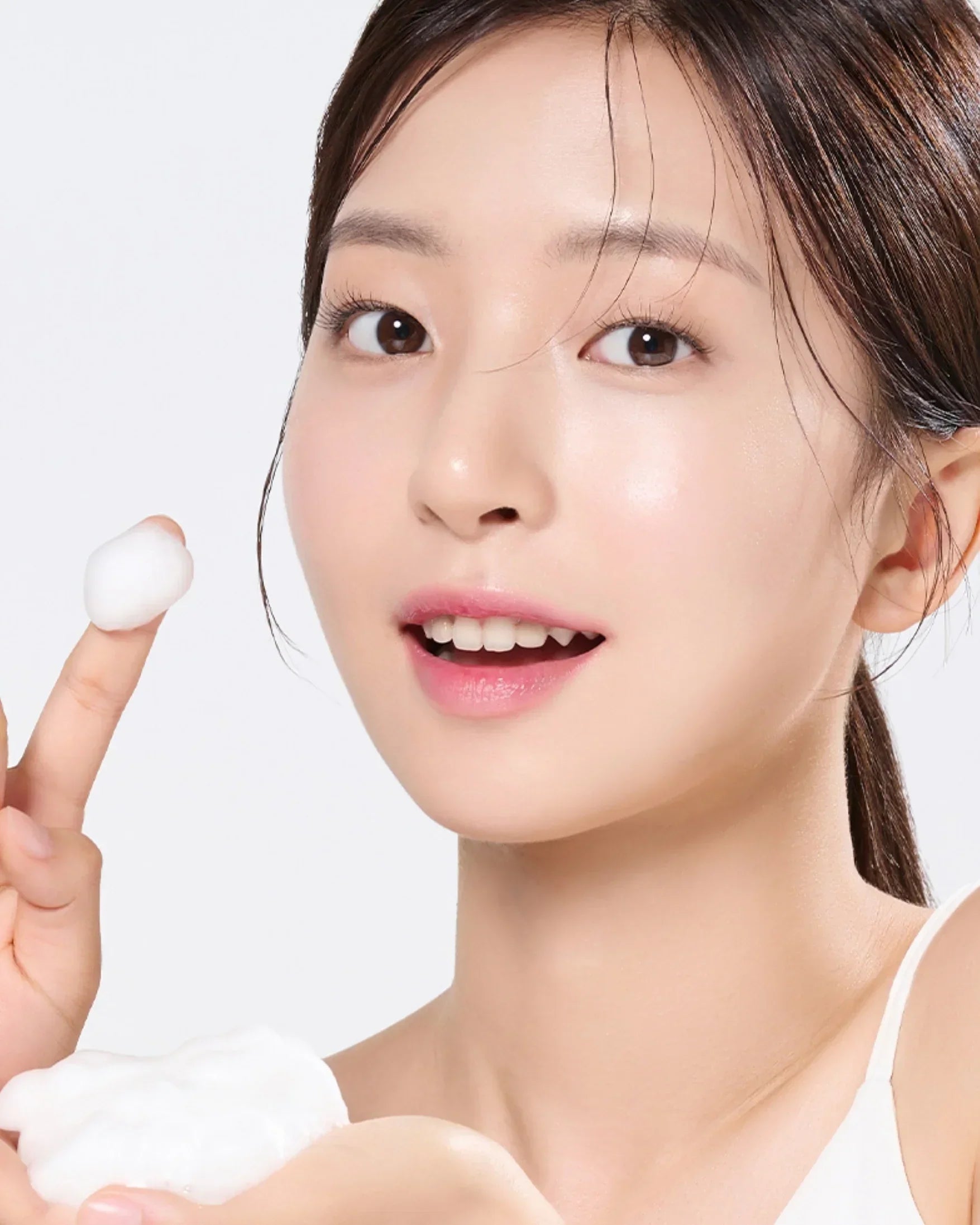 Young woman with glowing skin holding white foamy skincare product on finger against white background