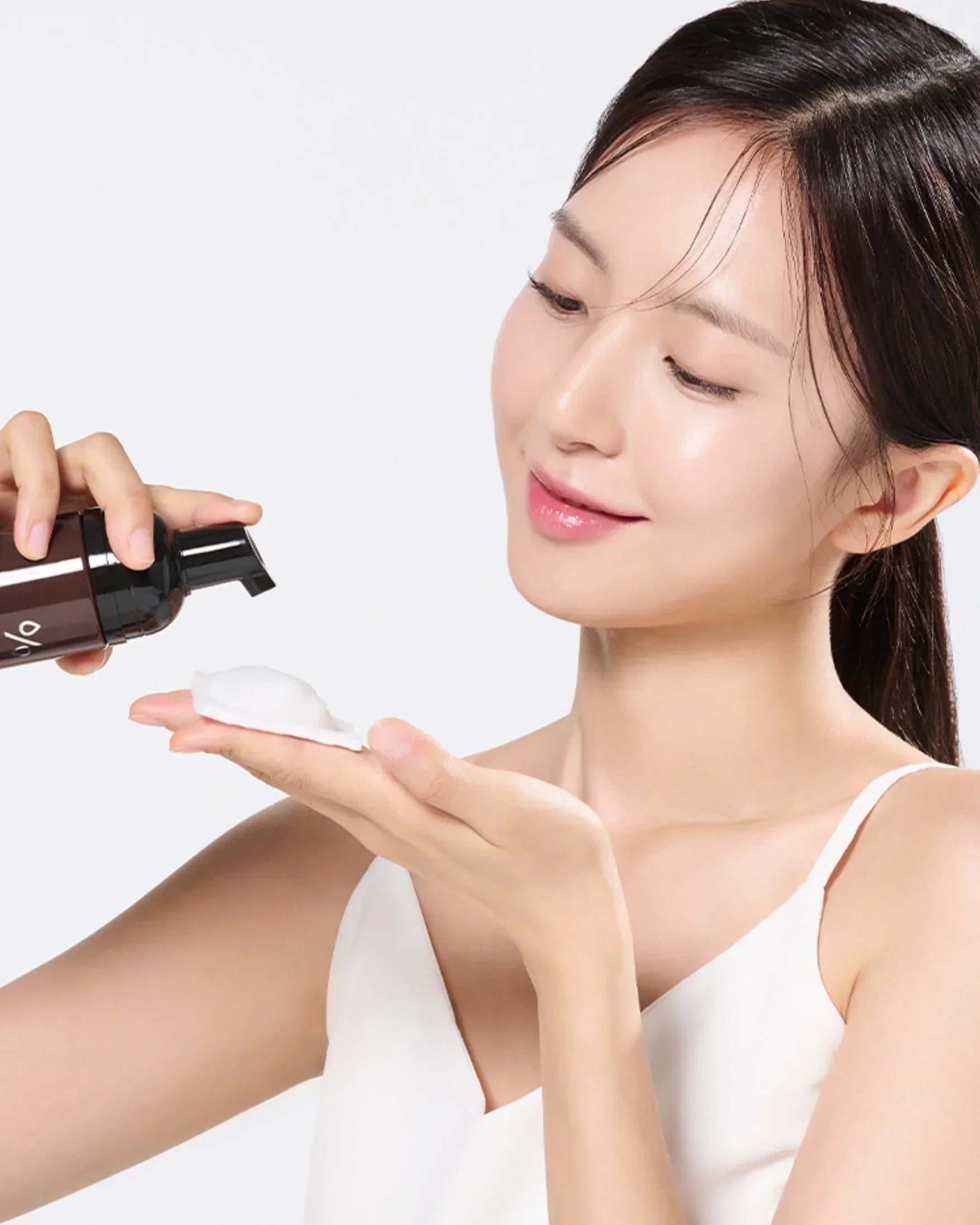 Woman with clear skin dispensing white foaming cleanser from a dark pump bottle into her hand