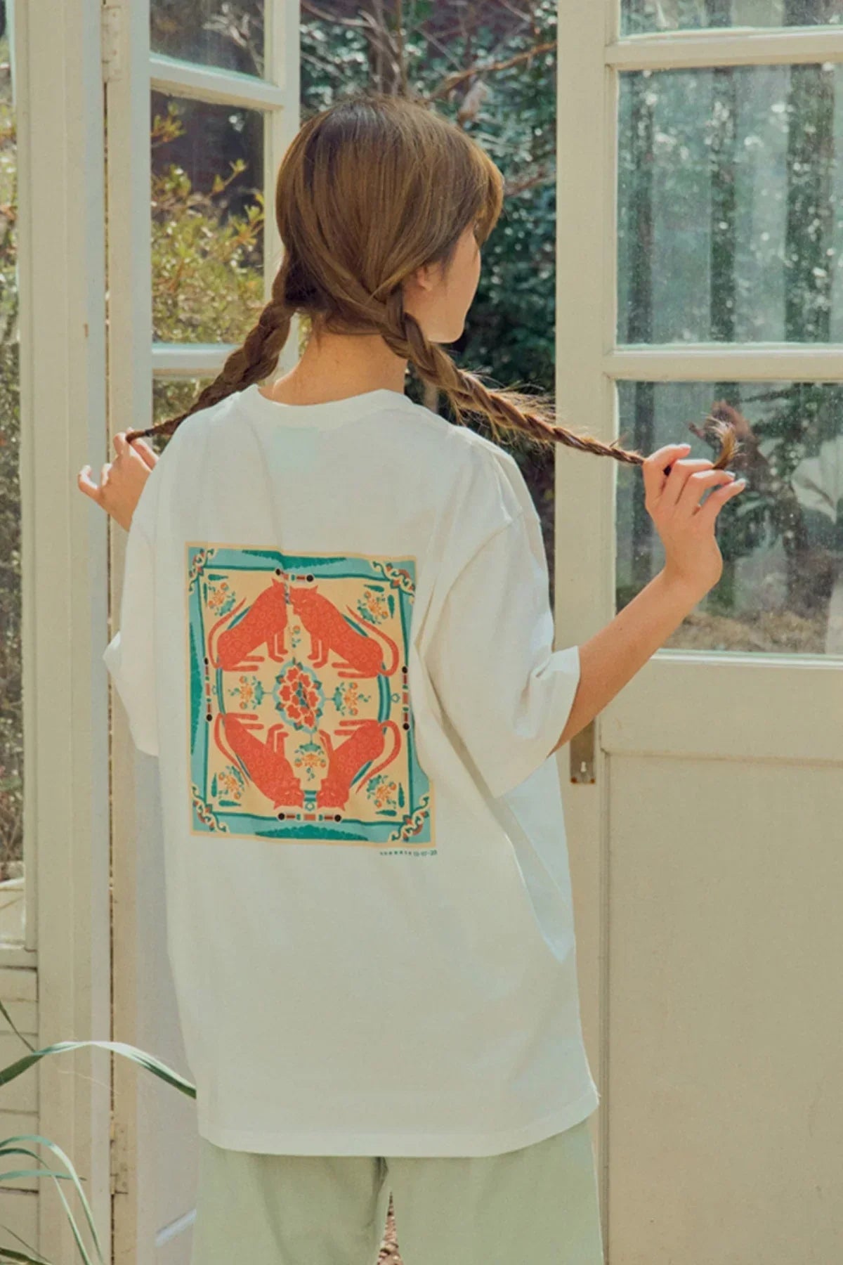 Woman indoors wearing white T-shirt with colorful red fox and floral back print, holding braided hair