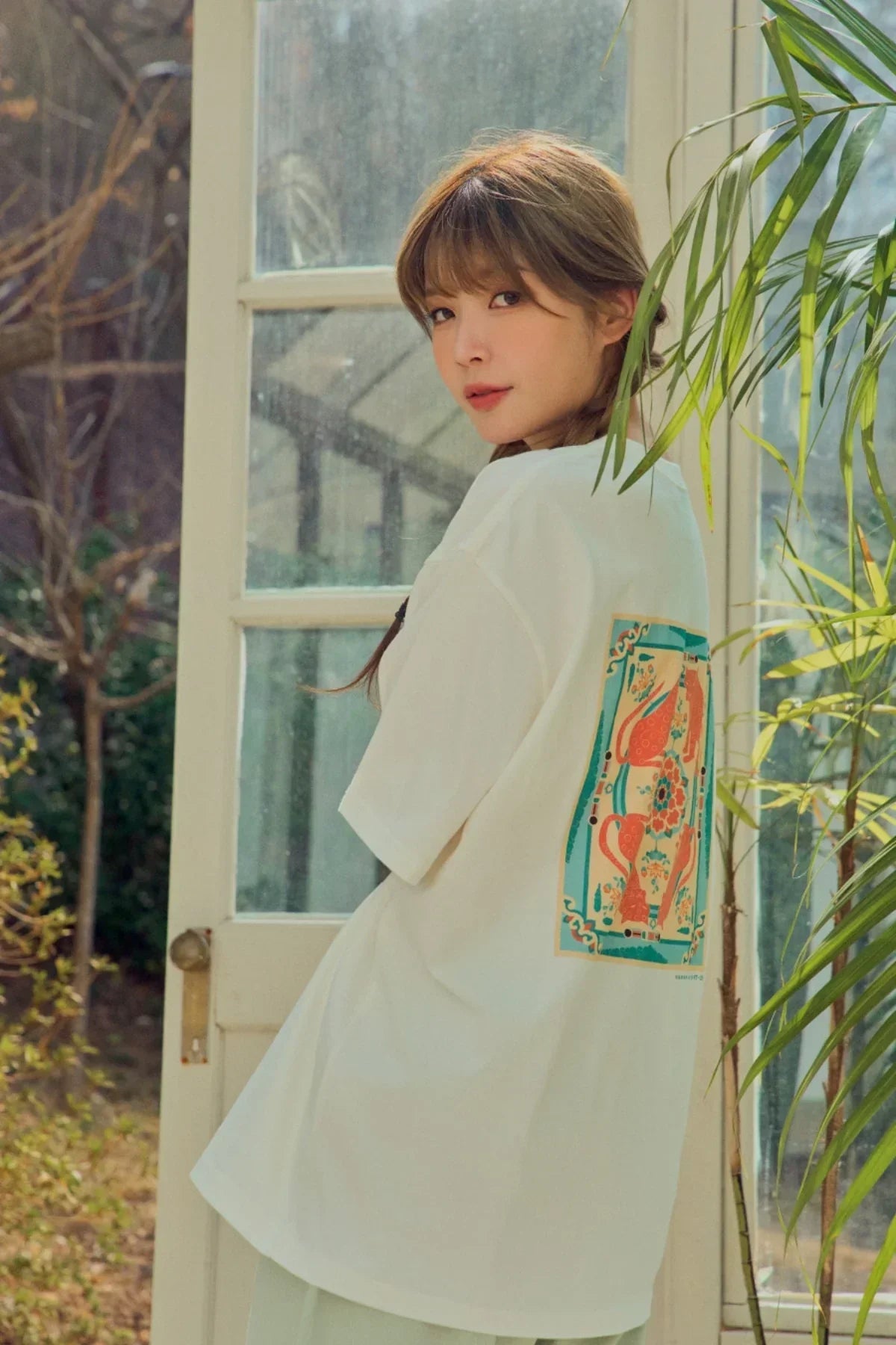 Woman wearing white T-shirt with colorful vintage vase print on back, standing indoors near window and plants