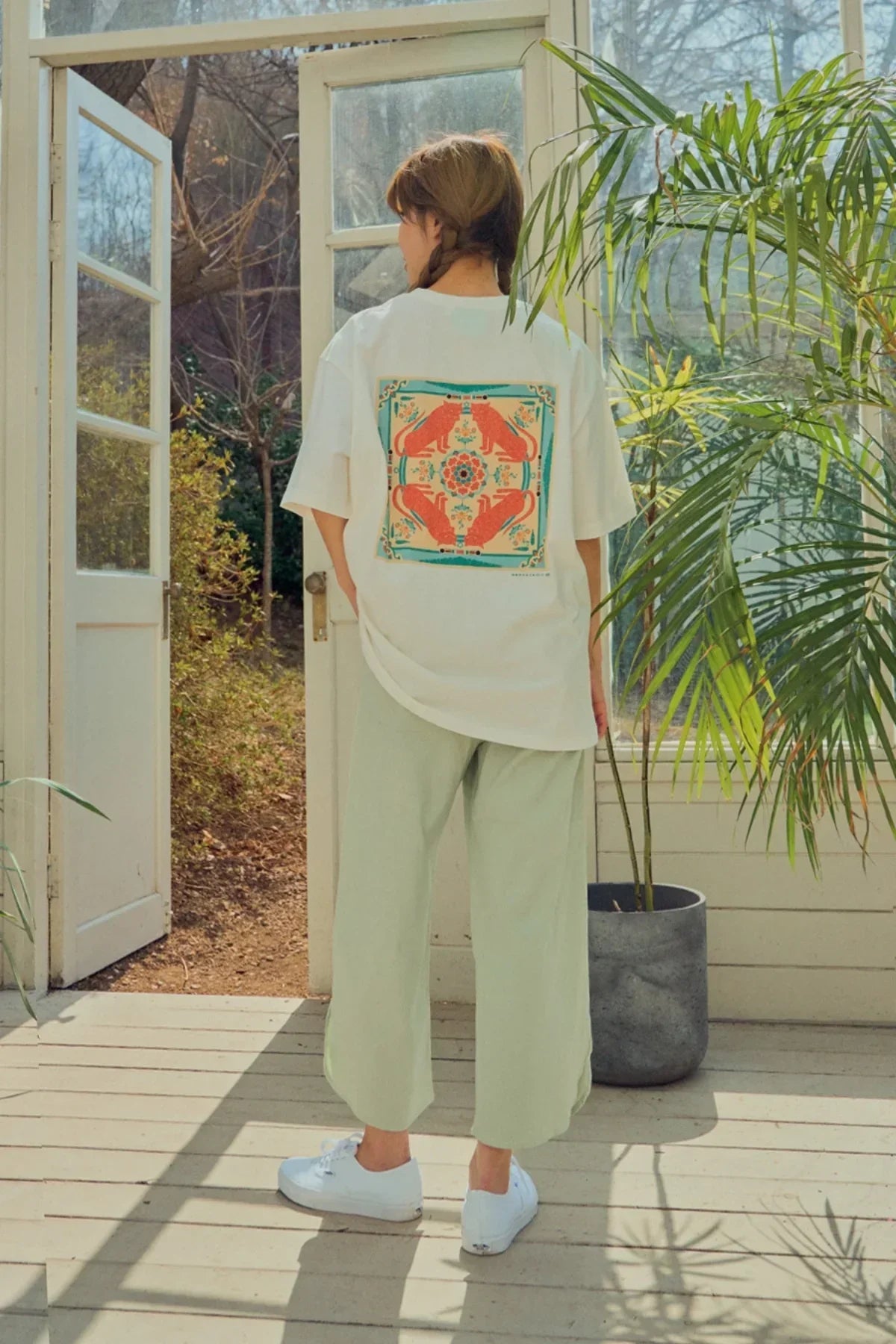 Woman wearing white graphic t-shirt with orange cat design, standing in sunlit room with plants
