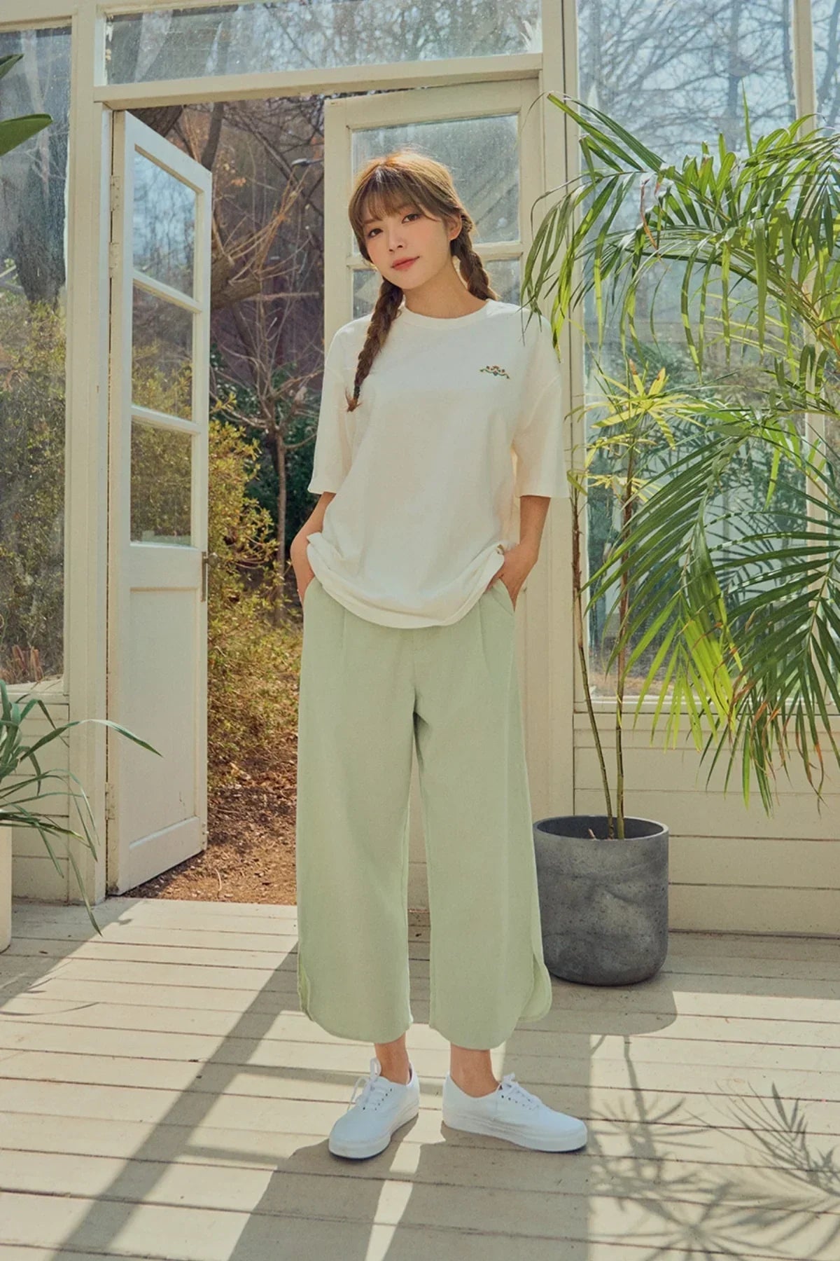 Young woman with braided hair wearing a white graphic t-shirt, light green wide pants, and white sneakers in a sunlit room with plants