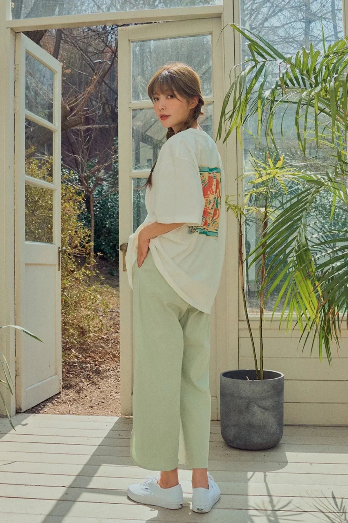 Woman in white graphic t-shirt and light green pants standing in sunlit open doorway with plants