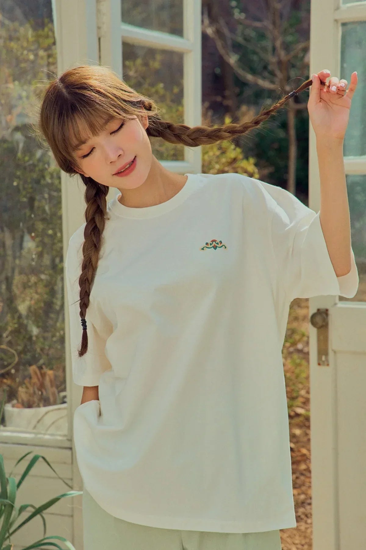 Young woman in a white oversized t-shirt with small colorful logo, playing with braided hair in sunlit room
