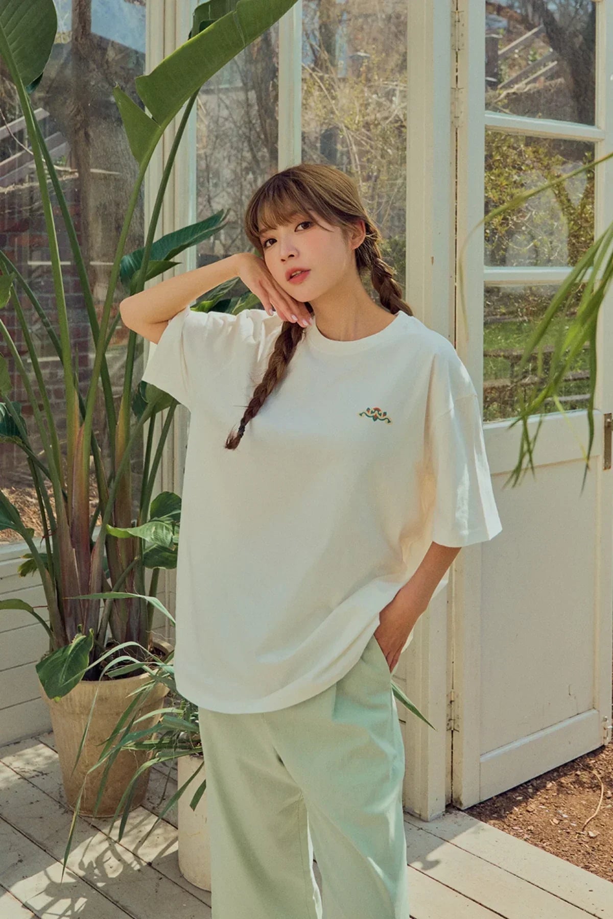 Woman wearing white oversized t-shirt with small snake embroidery, light green pants, standing in sunlit room with plants