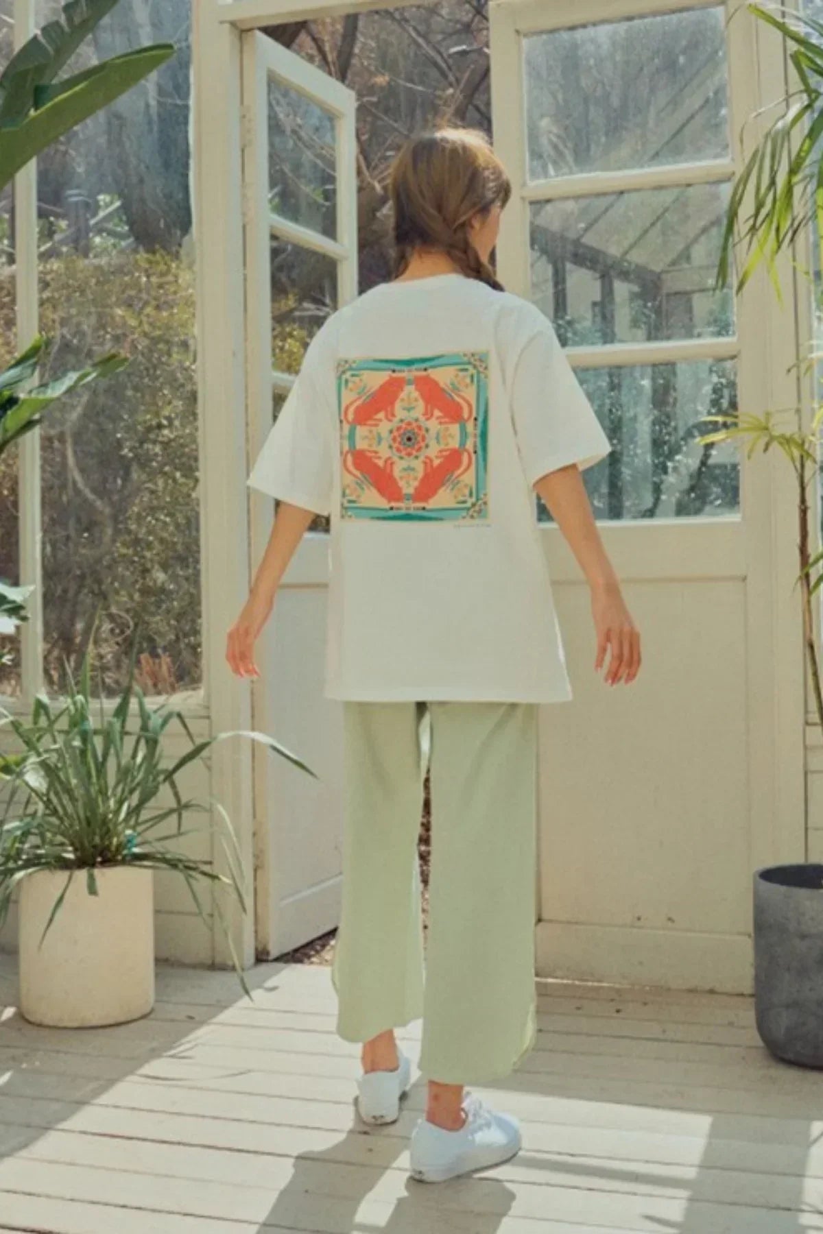 Woman standing in sunlit room wearing white t-shirt with colorful animal print on back, light green pants, white sneakers