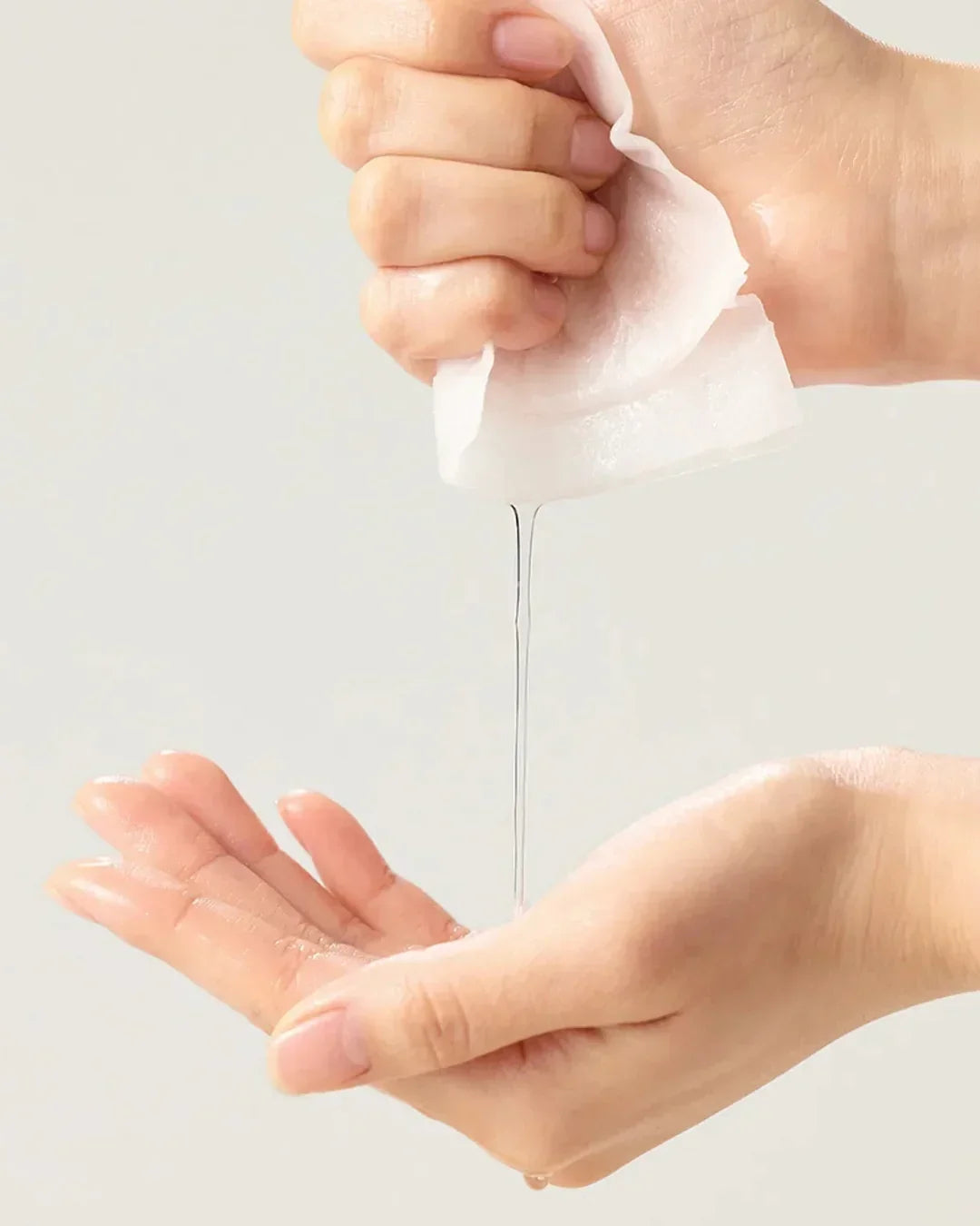 Hand squeezing liquid out of a wet white facial sheet mask over another open palm