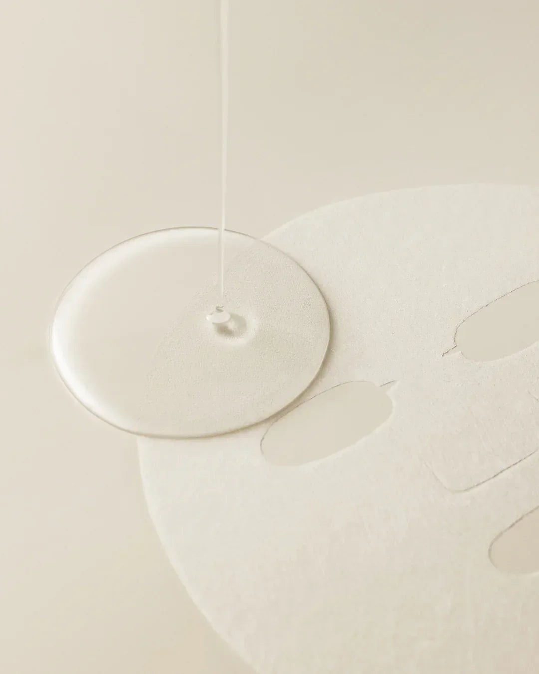 Skincare serum being poured onto a white textured sheet face mask on a beige background