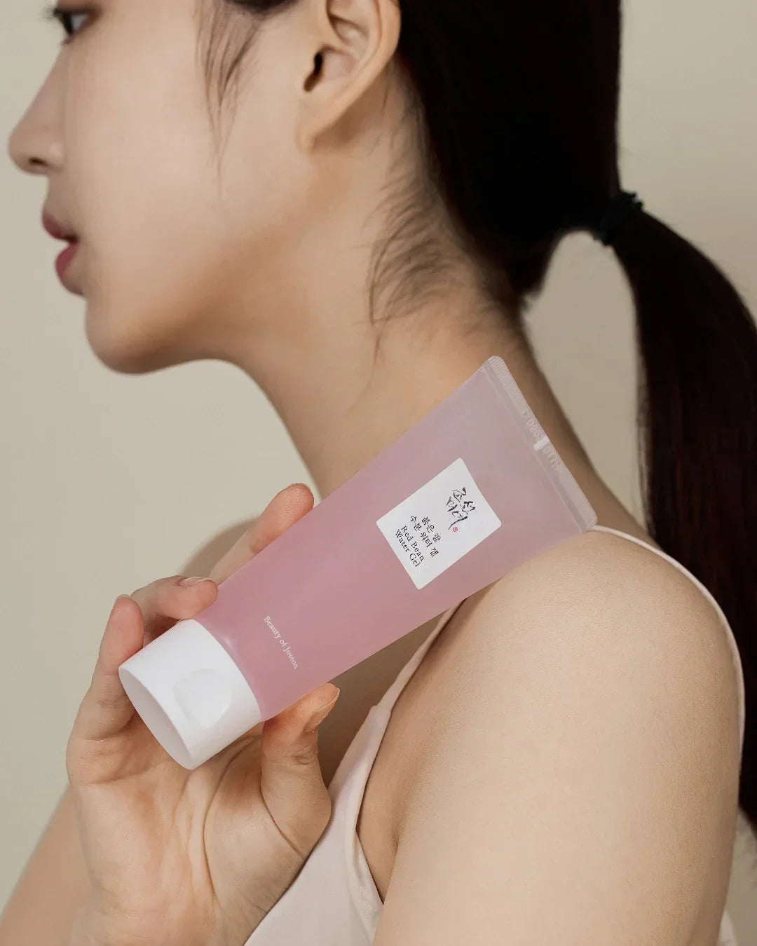 Woman holding pink tube of Red Bean Water Gel skincare product against beige background