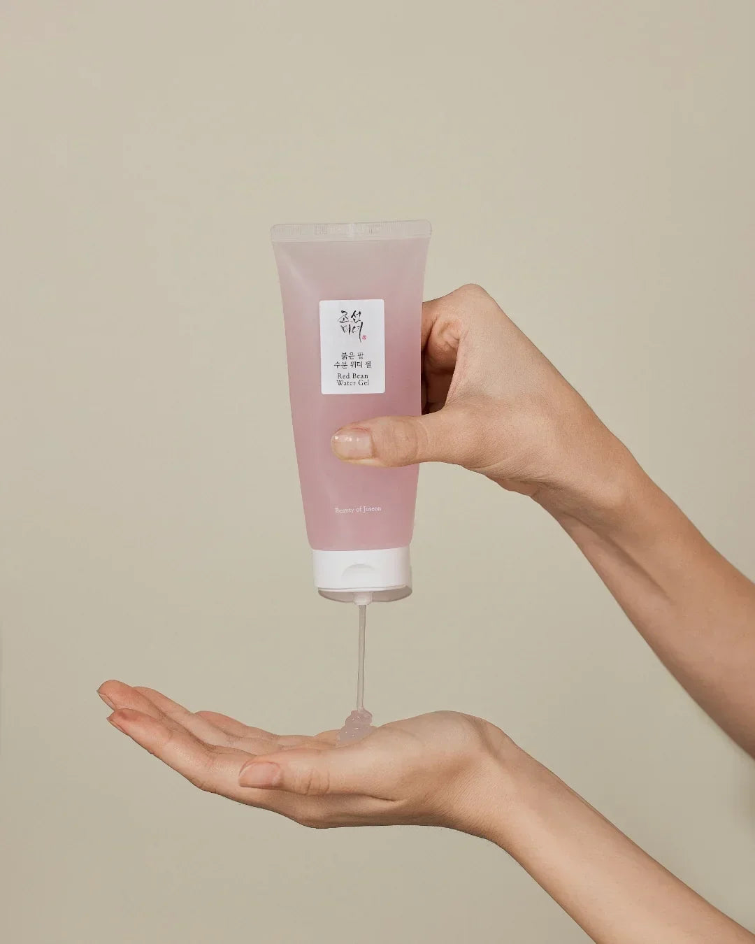 Hands dispensing Beauty of Joseon Red Bean Water Gel from pink tube on beige background